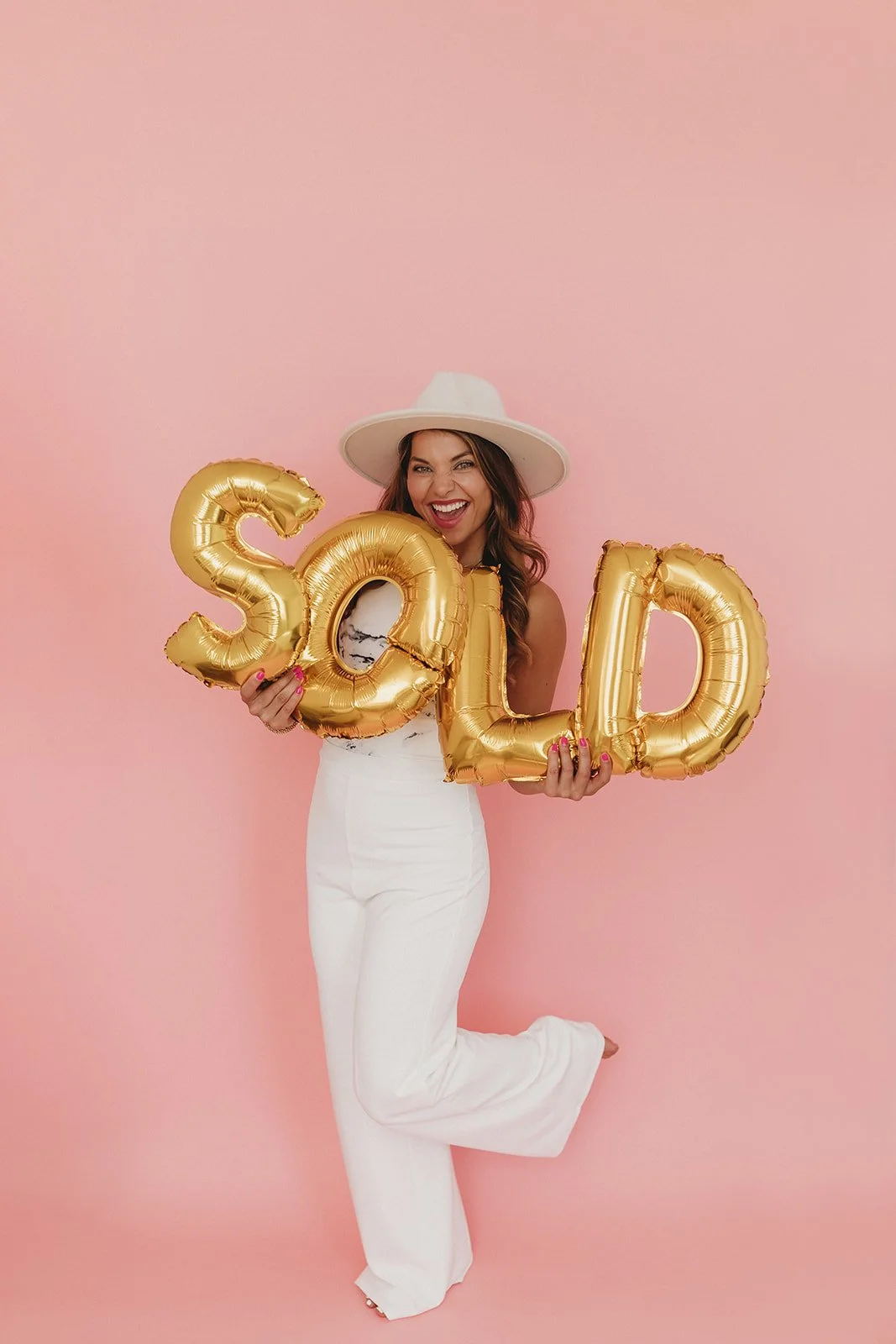 A woman wearing a white hat and white pants, holding gold balloon letters spelling "SOLD," standing against a pink background.