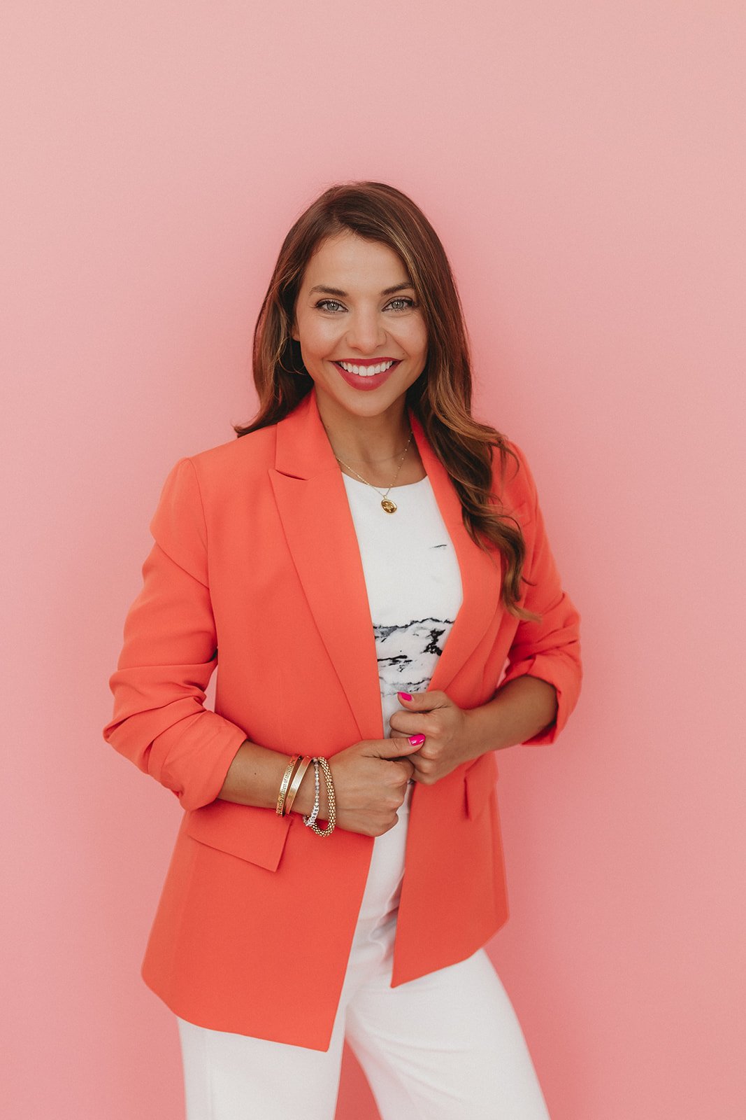 A smiling woman with long brown hair posing against a pink background, wearing a coral blazer over a white t-shirt, white pants, and multiple bracelets.