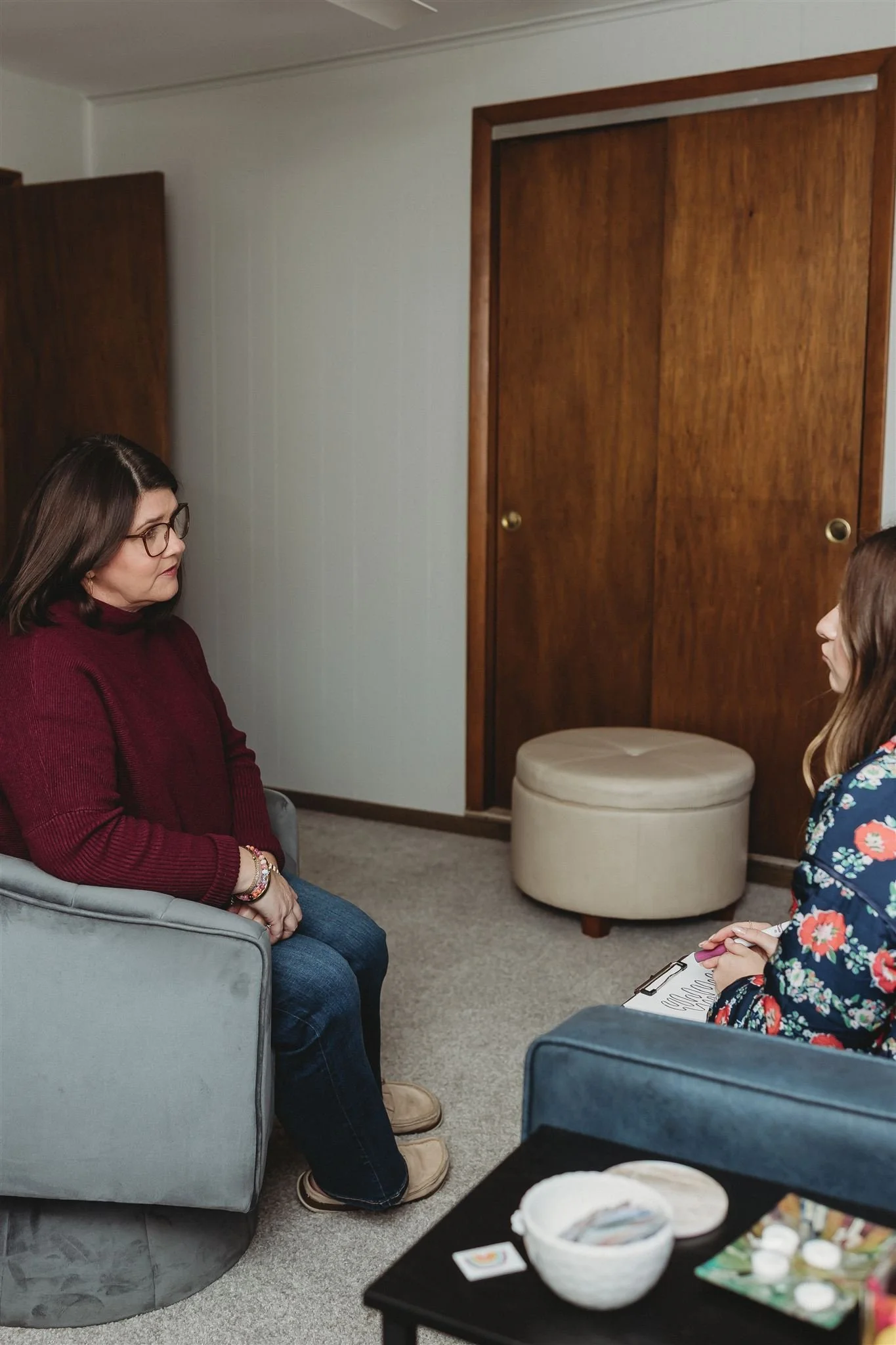 Two women sitting and talking inside a room, one on a gray chair and the other on a couch, with a wooden closet and a round ottoman behind them.