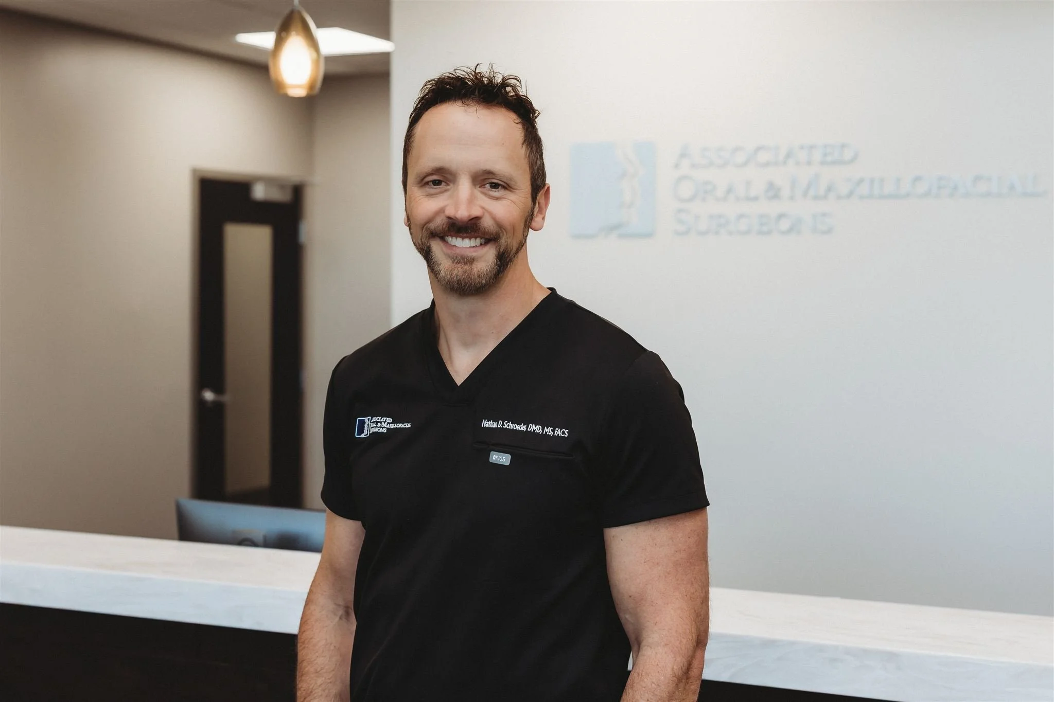 A smiling man in black medical scrubs standing in a modern office or clinic environment, with a blurred logo and text on the wall in the background.