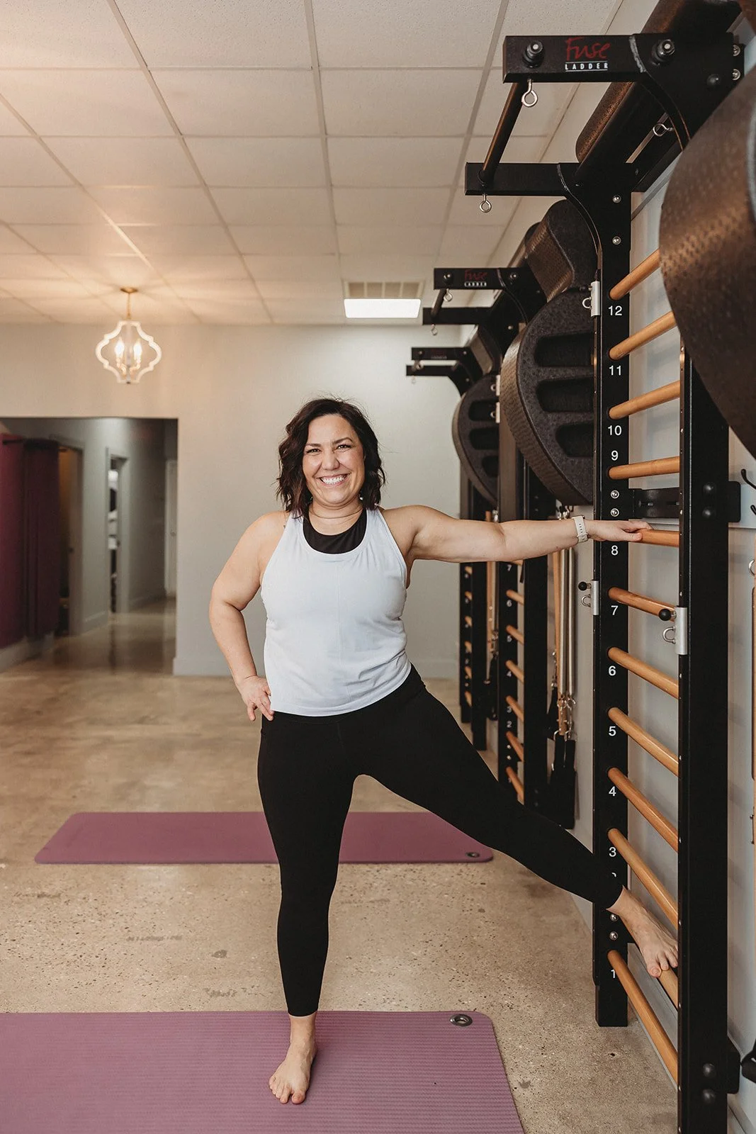 A woman smiling and stretching her arm while standing on a yoga mat in a fitness studio with a wall-mounted wall rack.