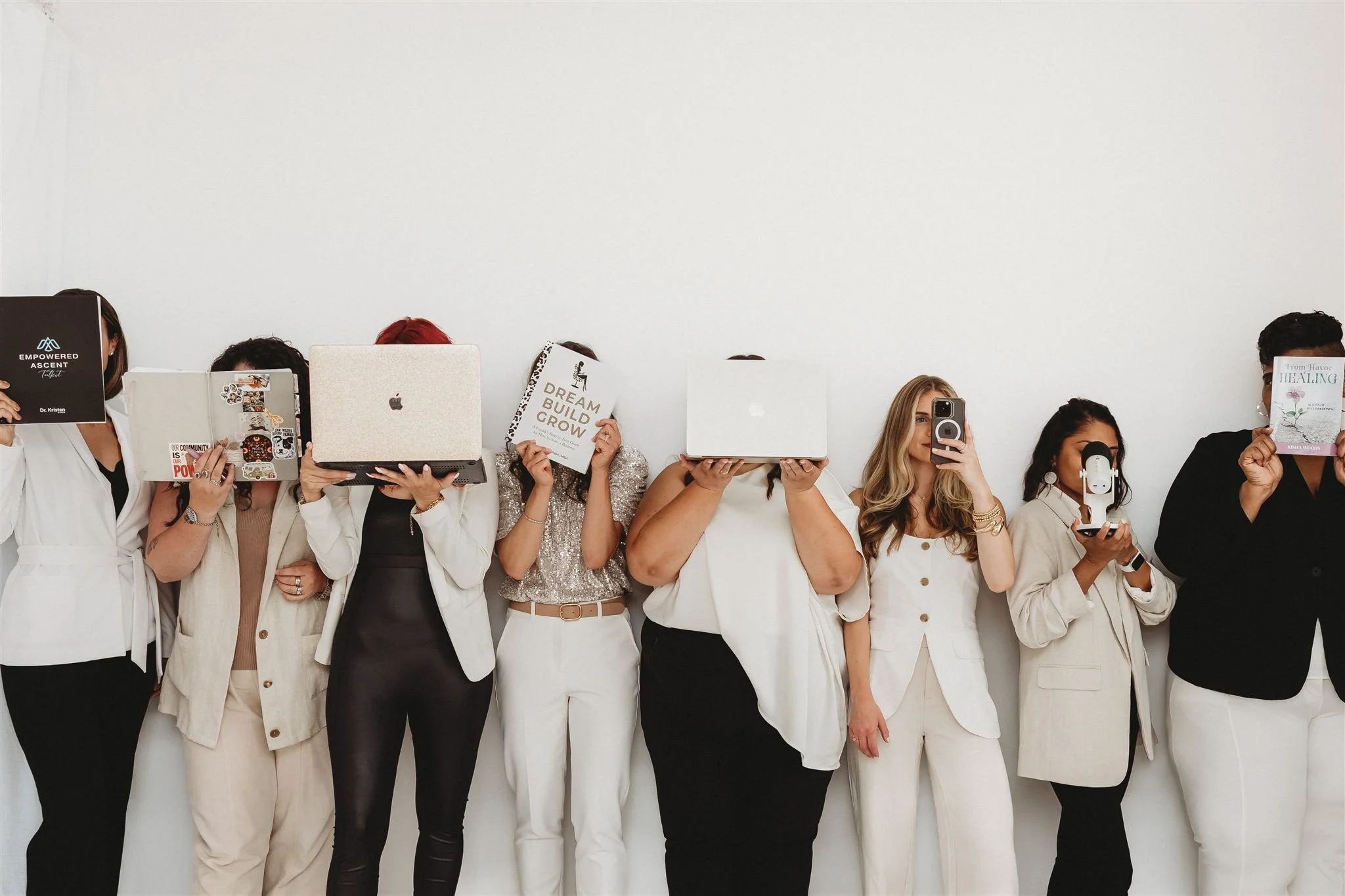 Group of women standing against a white wall, holding laptops and books in front of their faces, with some also using phones.