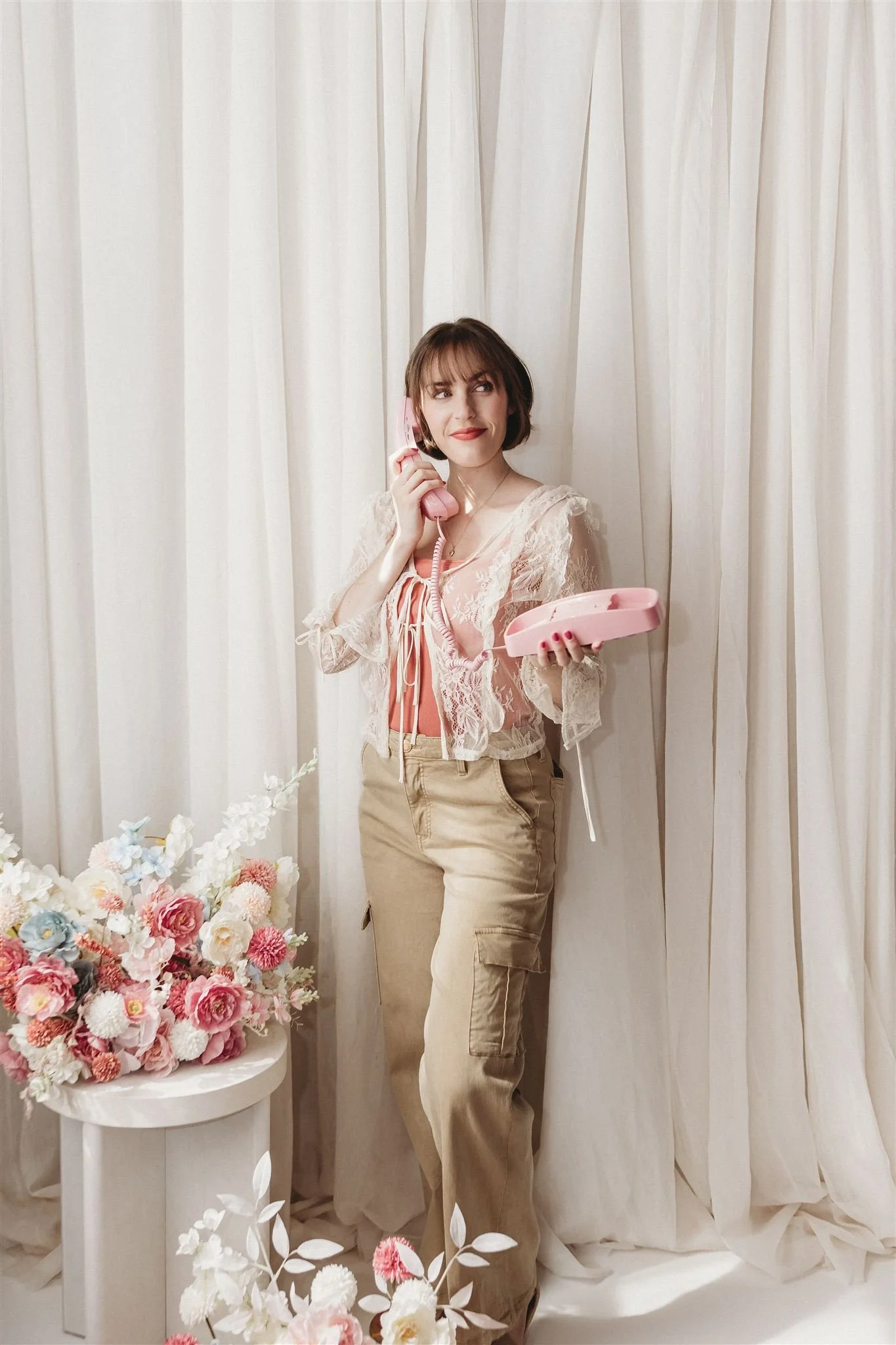 A young woman with short brown hair stands in front of cream-colored curtains, holding a pink telephone handset to her ear while looking slightly to the side with a smile. She is wearing beige cargo pants and a pink top with a sheer, lace cropped jacket. There is a white table with a large bouquet of pink, white, and blue flowers in front of her.