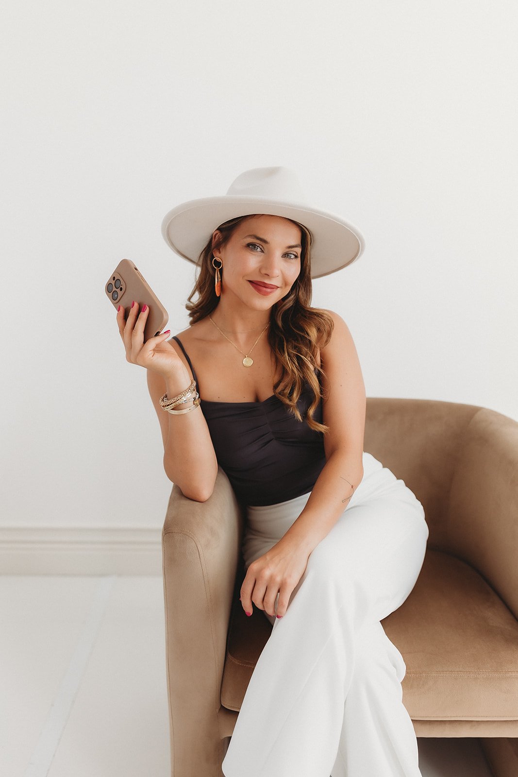 A woman with long wavy brown hair, wearing a large white hat, a black tank top, and white pants, sits on a beige sofa holding a smartphone and smiling at the camera.