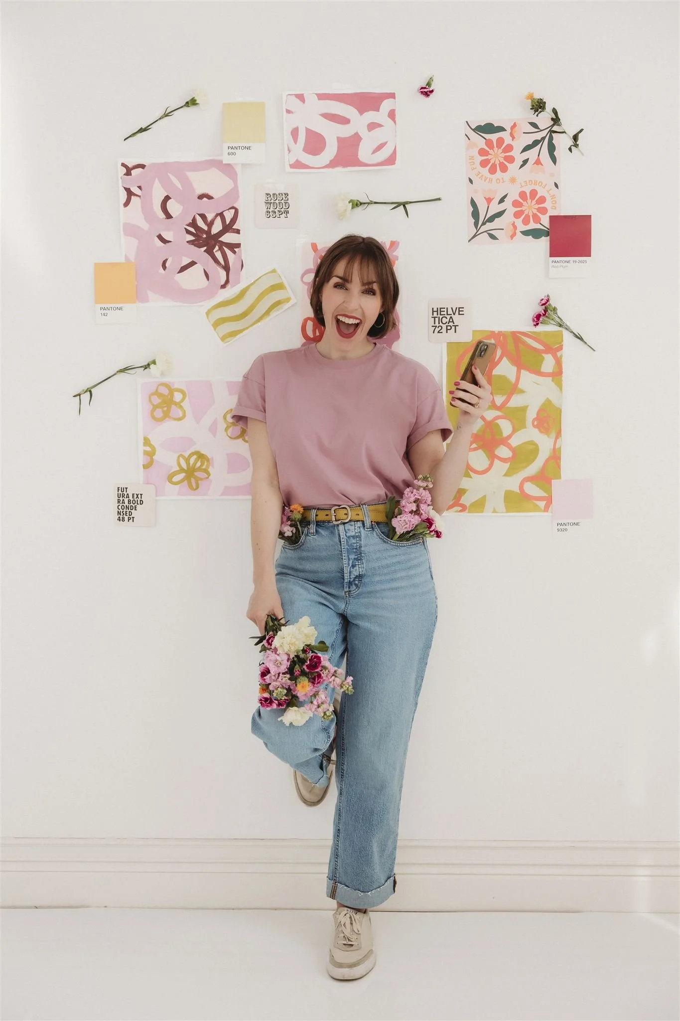 A young woman standing in front of a white wall decorated with colorful abstract art pieces and flowers, smiling and holding a phone in one hand and a bouquet of flowers in the other.