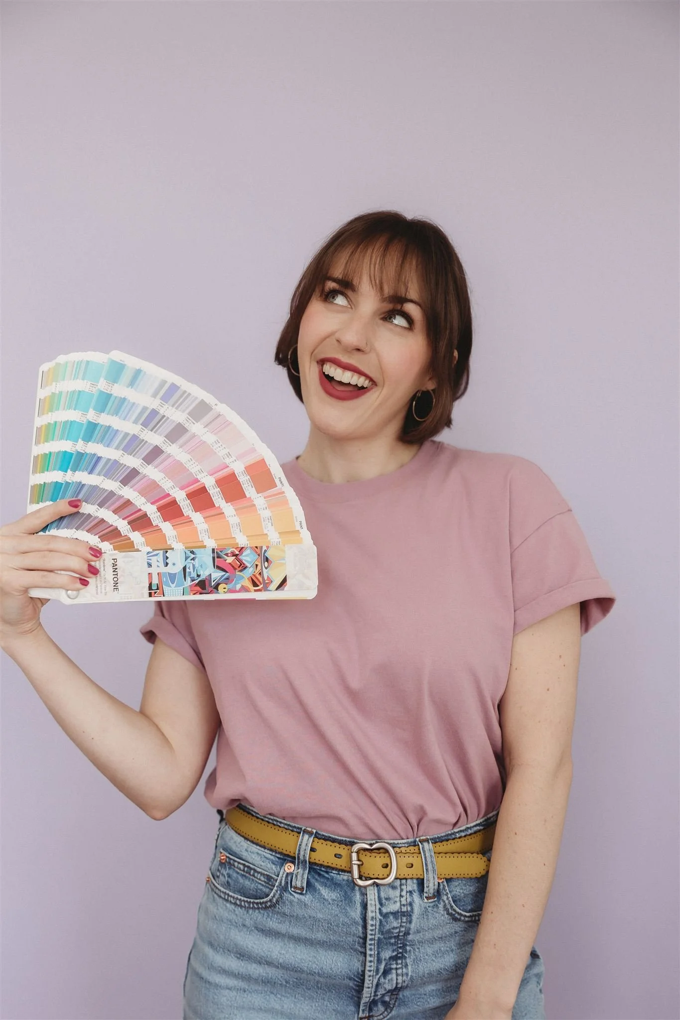 A woman with short brown hair, wearing a pink t-shirt and blue jeans, is holding a color fan with various colors and smiling happily.