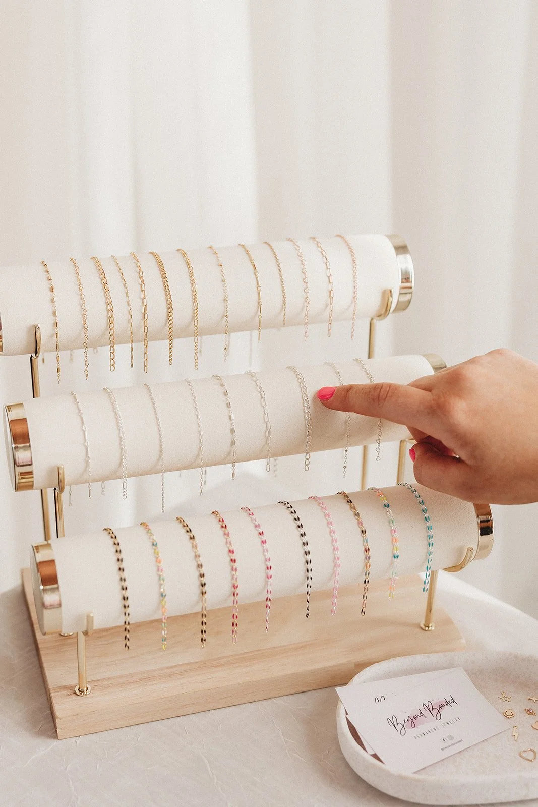 Four horizontal display racks of colorful bracelets and necklaces, with a person's hand pointing to one of the bracelets on the second rack, and a small tray with jewelry-making notes and small pouches in the foreground.