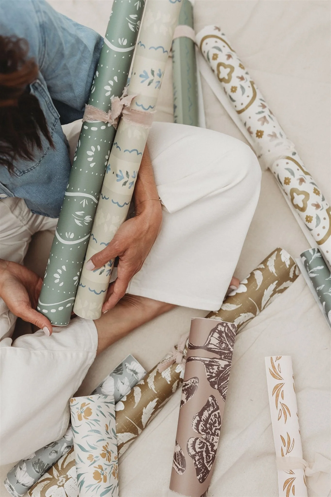 Person wrapping floral and patterned gift paper with various rolls of wrapping paper around.