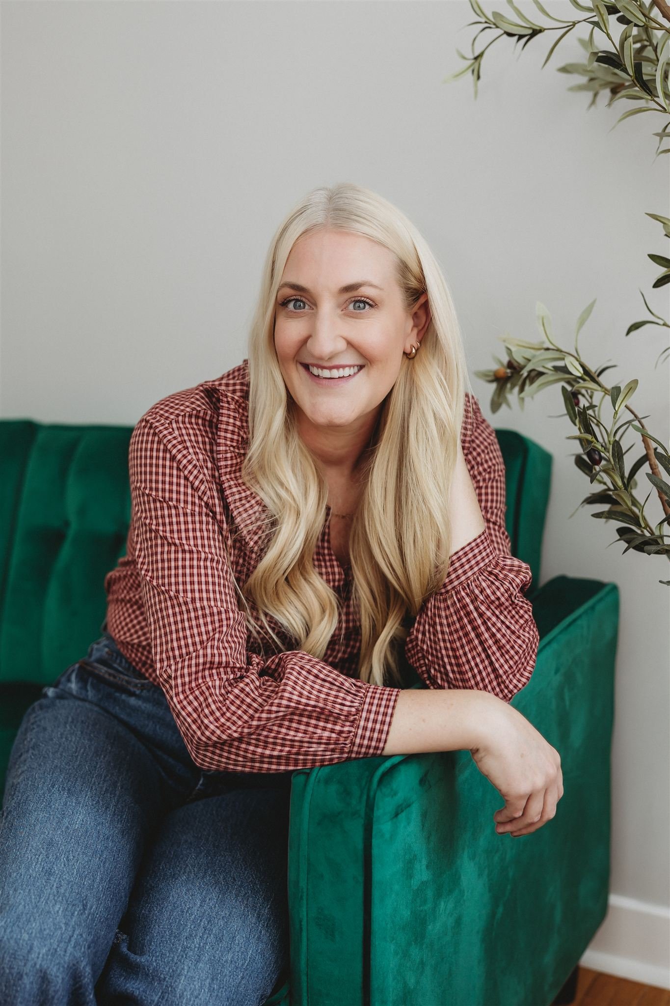 A smiling blonde woman sitting on a green velvet couch, wearing a red checkered shirt and blue jeans, with a plant on the right side of the frame and a plain light-colored wall in the background.