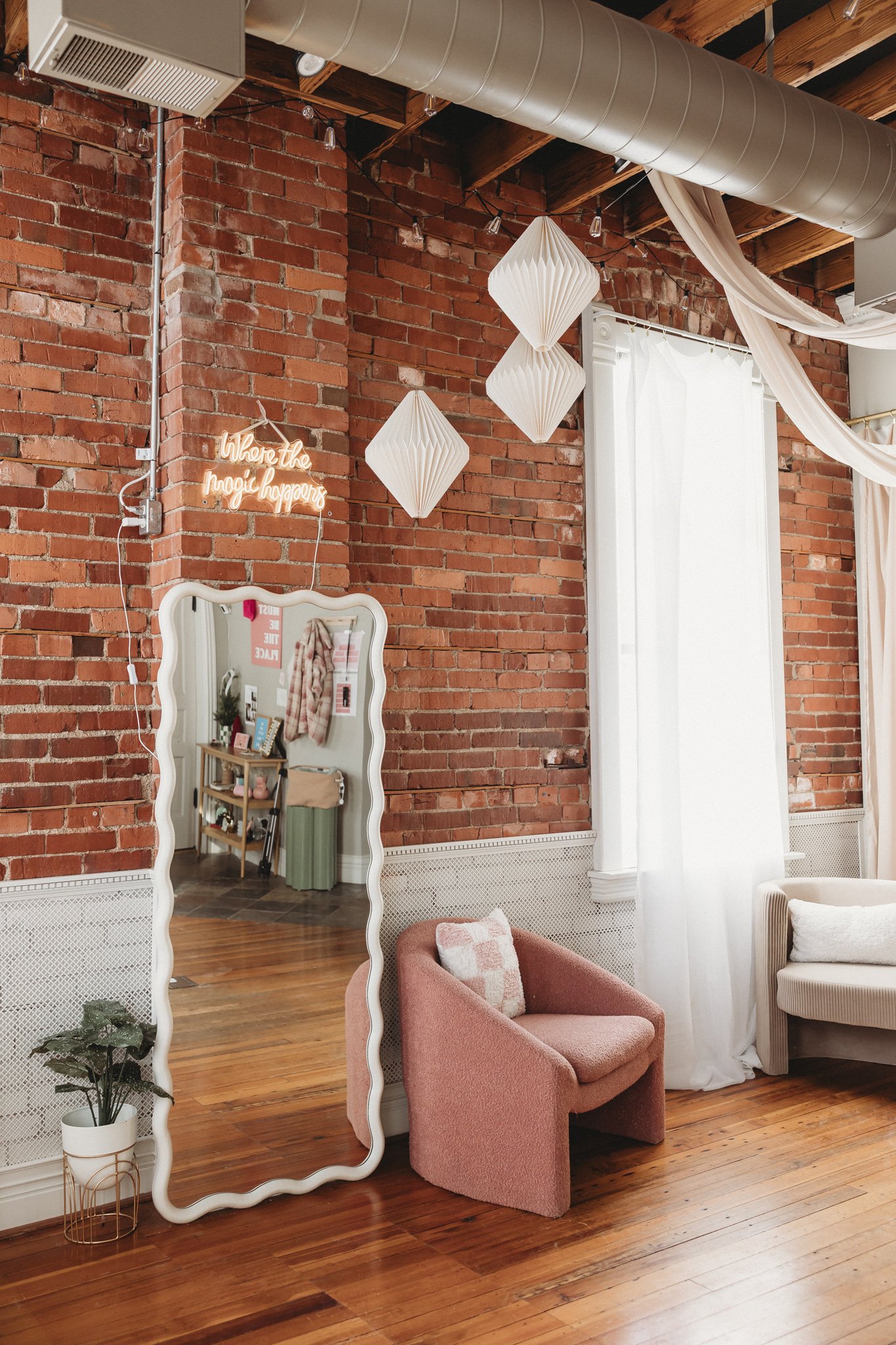 Interior of a room with a brick wall, white curtains, a large mirror with a wavy frame, a small pink armchair with a patterned pillow, a white sofa, hanging white paper lanterns, a neon sign that reads 'Where the magic happens,' and some decorative elements.