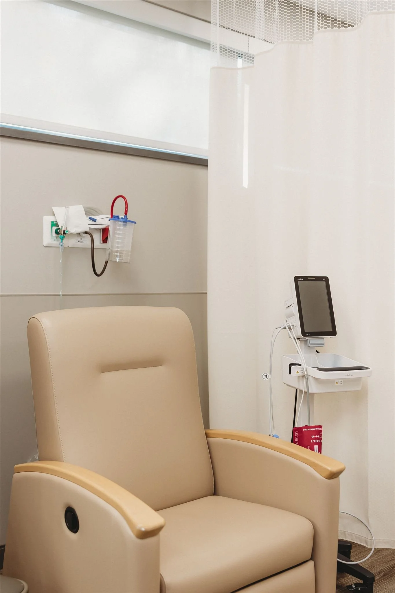 Empty hospital or clinic chair with a medical monitor or screen attached to the wall nearby, and medical equipment mounted on the wall, including a waste container and tubing.