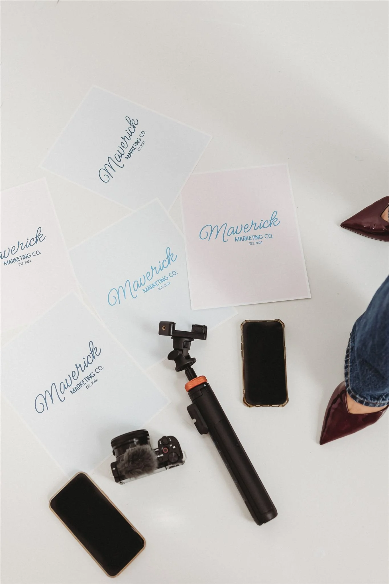 Flat lay of Maverick Marketing Co. branding materials, two smartphones, a camera with microphone, a tripod, and a person's legs wearing jeans and maroon boots.