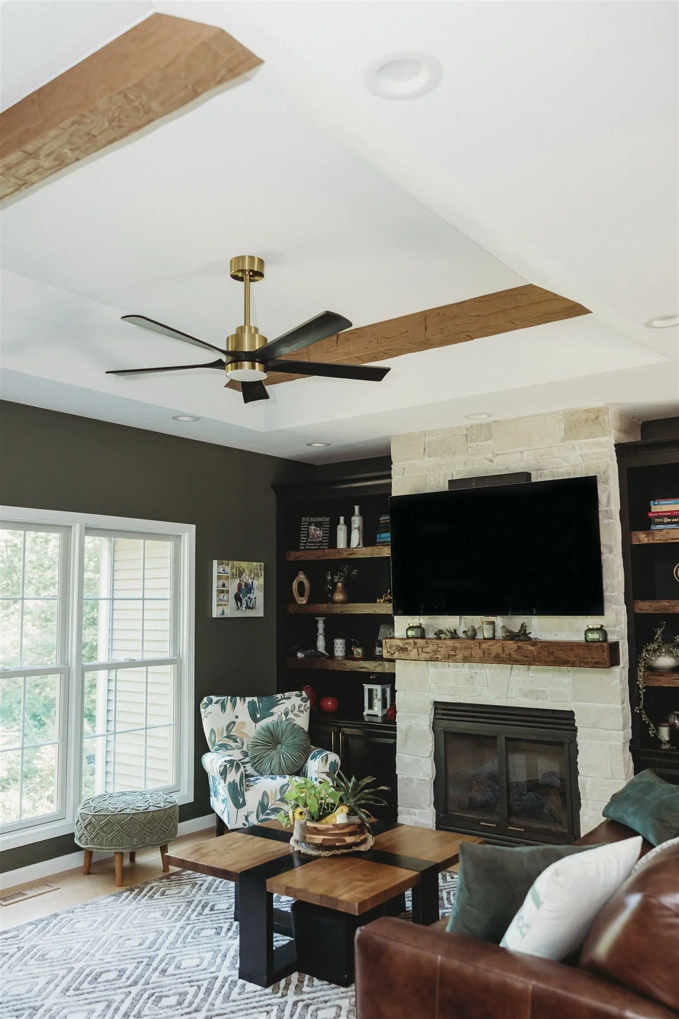 Living room with a ceiling fan, large window, armchair with a blue floral pattern, a small ottoman, a wooden coffee table with a plant, a stone fireplace with a mounted TV, and built-in dark wood shelves filled with decor.