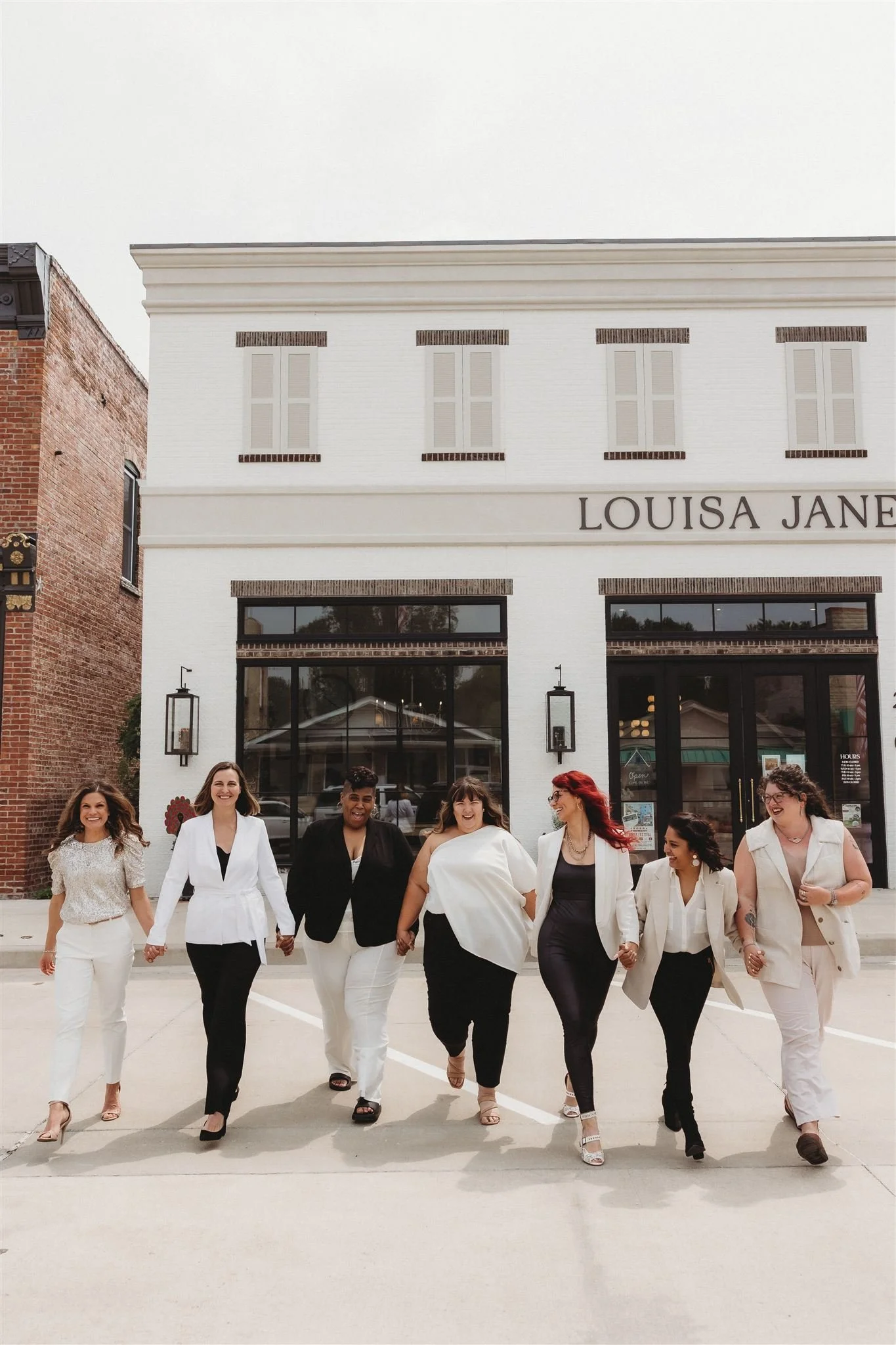 Seven diverse women walking and holding hands outside of a white storefront with the sign "Louisa Jane" on it.