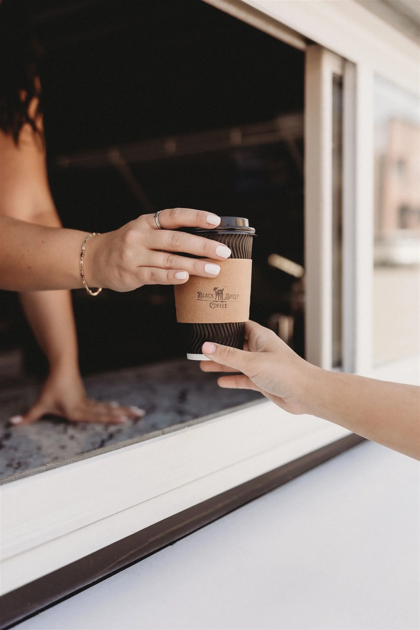 A person is handing a paper cup of coffee to another person through a window, with the cup labeled 'Black Sheep Coffee'