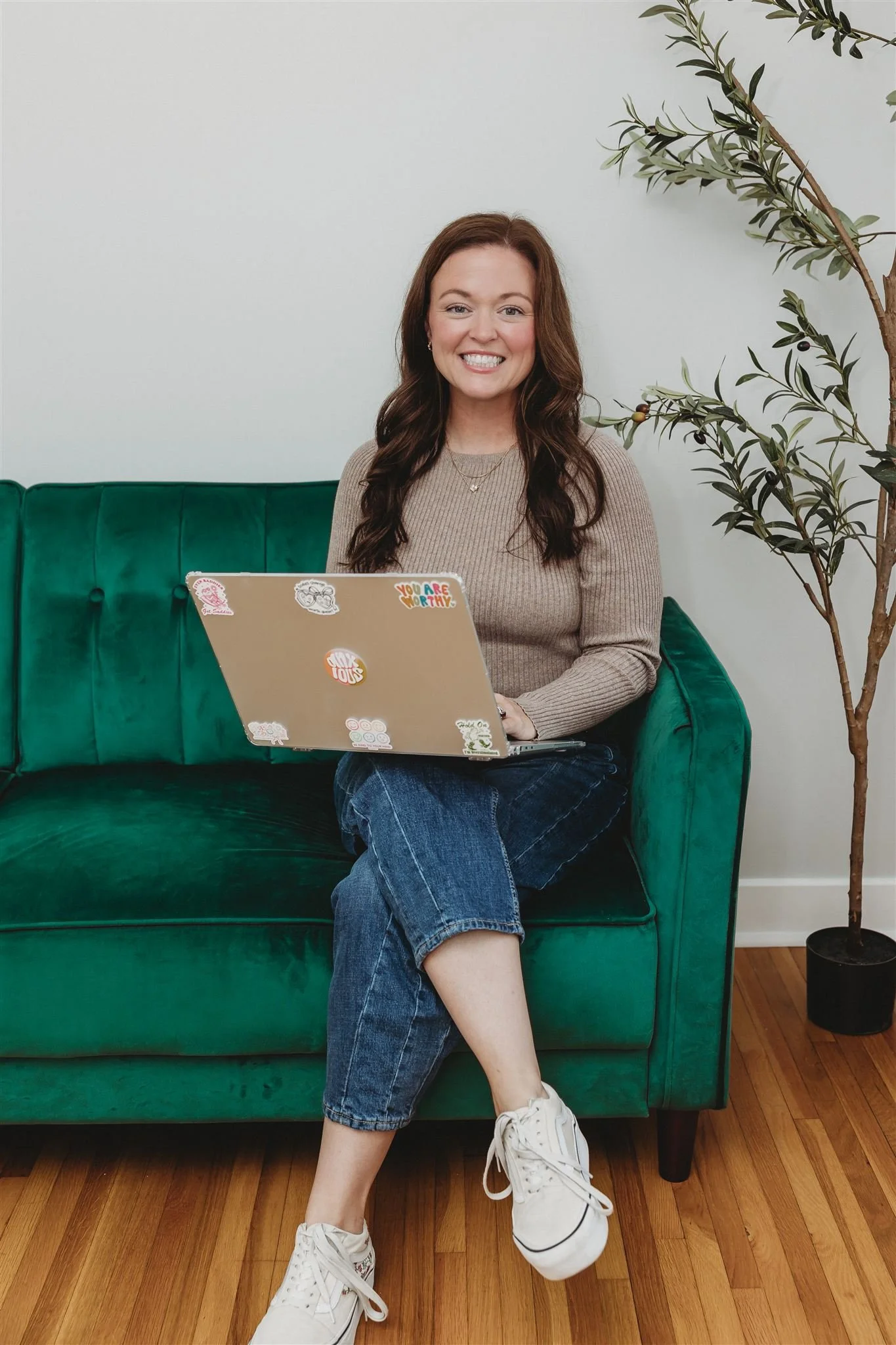 A woman with long brown hair sitting on a green velvet sofa, smiling, holding a laptop with stickers, wearing a beige sweater, blue jeans, and white sneakers, with a potted plant beside her on a wooden floor.