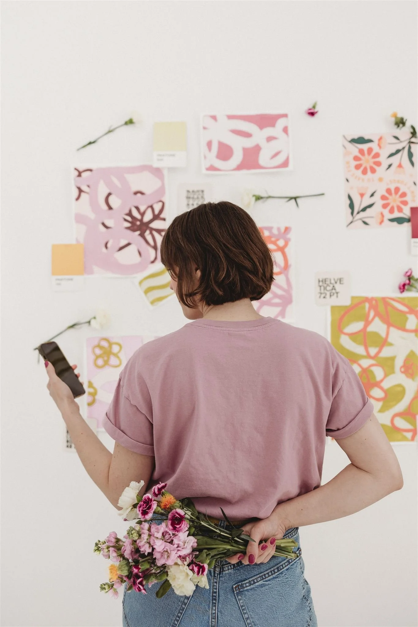 A woman with short brown hair wearing a pink T-shirt and jeans, holding a bouquet of pink, white, and purple flowers behind her back while looking at her phone in front of a wall decorated with colorful abstract art and flowers.