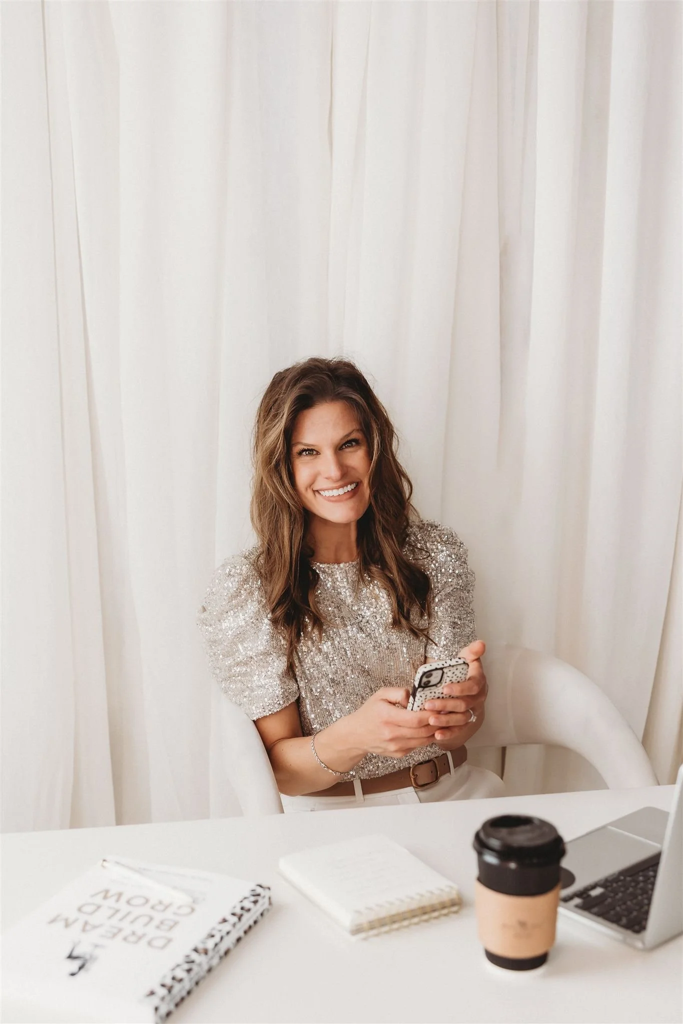A smiling woman with long wavy hair wearing a sparkly silver top, sitting at a white desk with a laptop, notebook, and coffee cup, in front of cream-colored curtains.