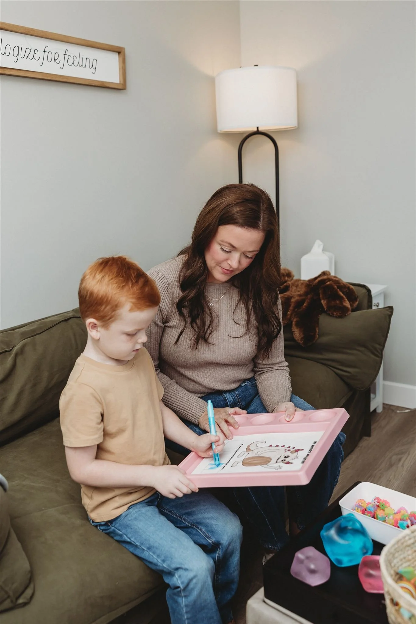 A woman and a young boy sitting on a green couch, looking at a pink tray with a drawing of a dog on it. The boy is holding a blue marker, and they appear to be coloring. Behind them is a floor lamp, a tissue box, and a plush brown dog on the couch.