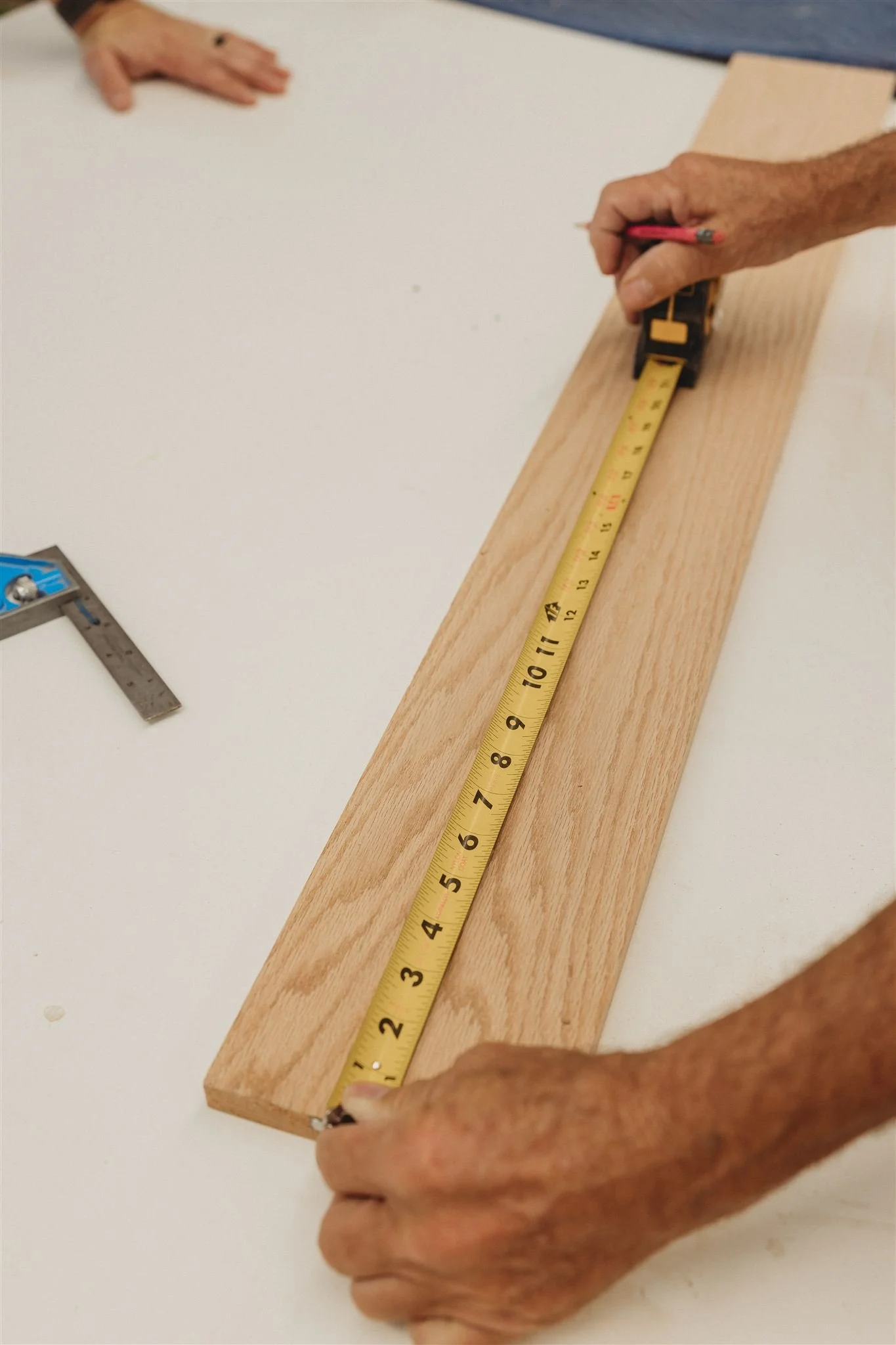 Two people measuring a piece of wood with tape measure, one person’s hand holding the tape and the other person’s hand is on the tape measure, not visible.