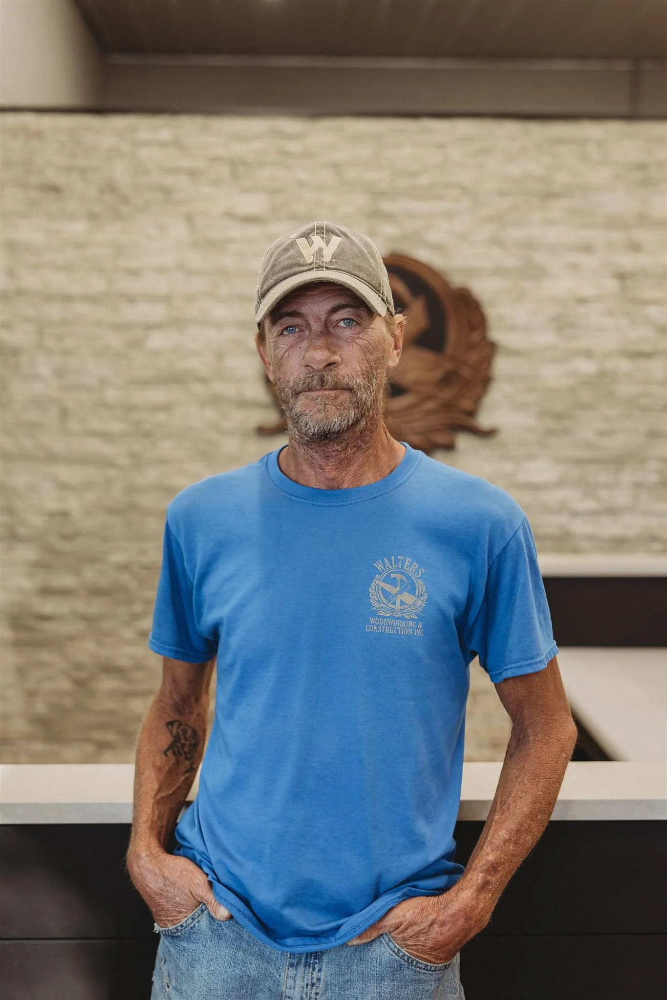 A rugged man with tattoos on his forearms, wearing a blue t-shirt and a gray baseball cap, stands indoors against a brick wall with a wooden carving behind him.