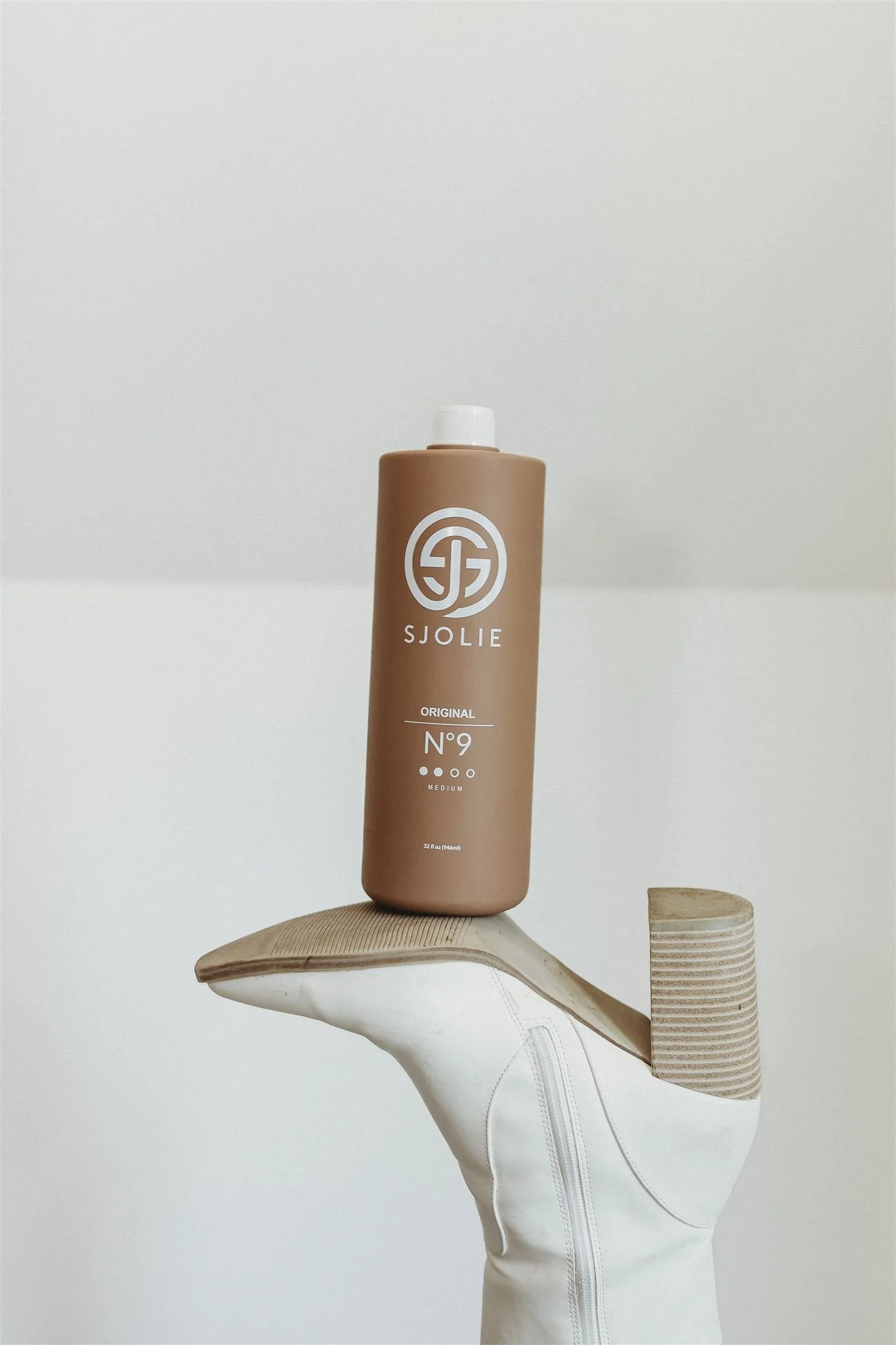 A beige shoe sole is balanced on a white mannequin foot, supporting a beige bottle of SJOLIE hair product, against a plain white background.