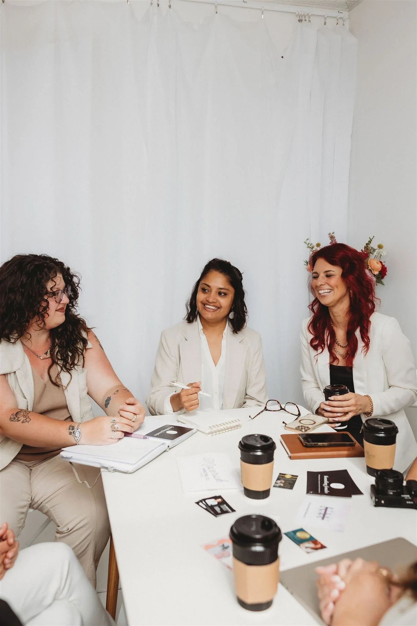 Three diverse women smiling and talking at a white table with coffee cups, notes, and a smartphone during a meeting or discussion.