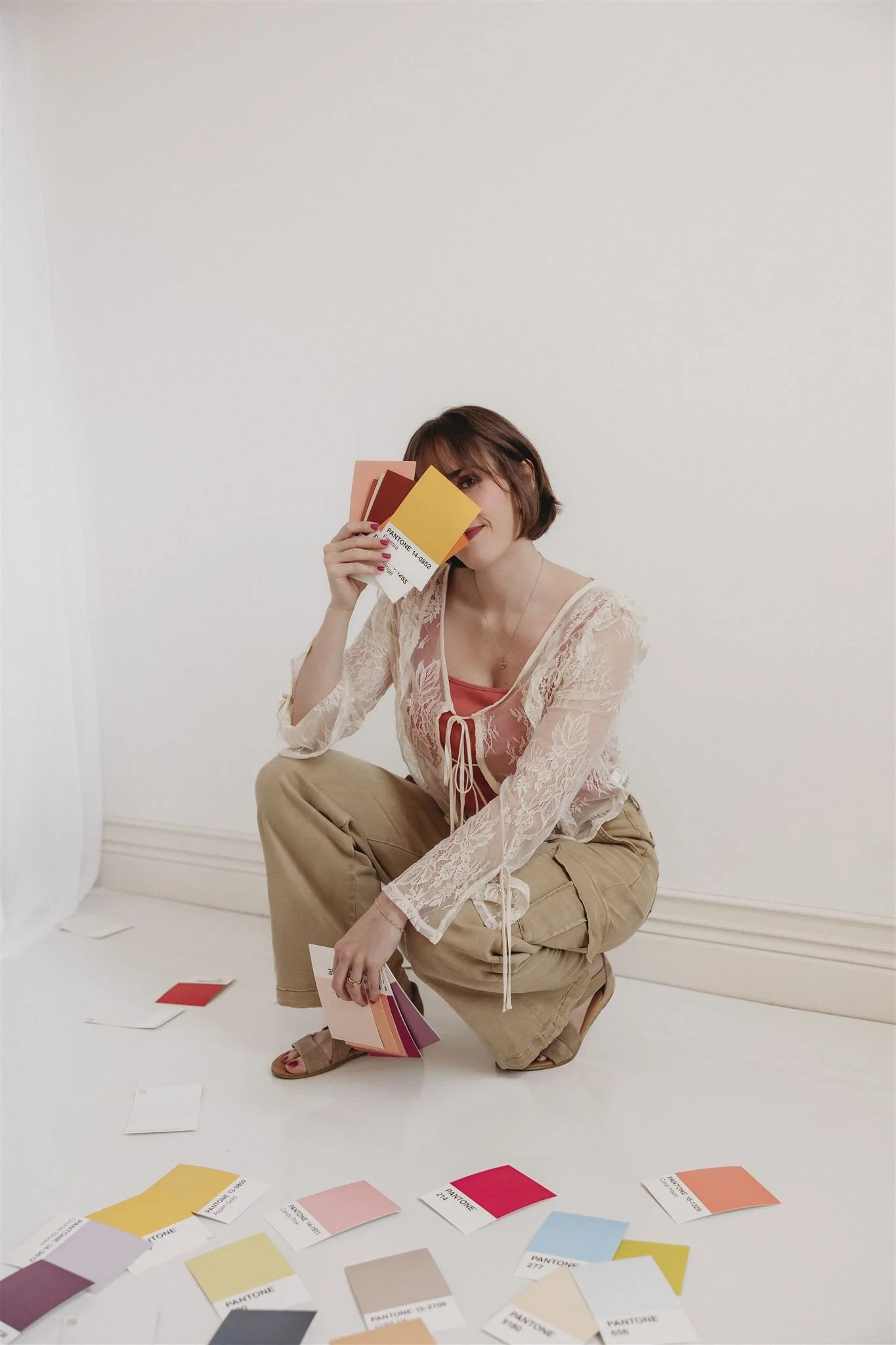 A woman with short brown hair is squatting on the floor in front of a plain white wall, holding up color swatches in front of her face. She is wearing a sheer lace long-sleeve top over a pink camisole, beige cargo pants, and sandals. Several color swatches are scattered on the floor around her.
