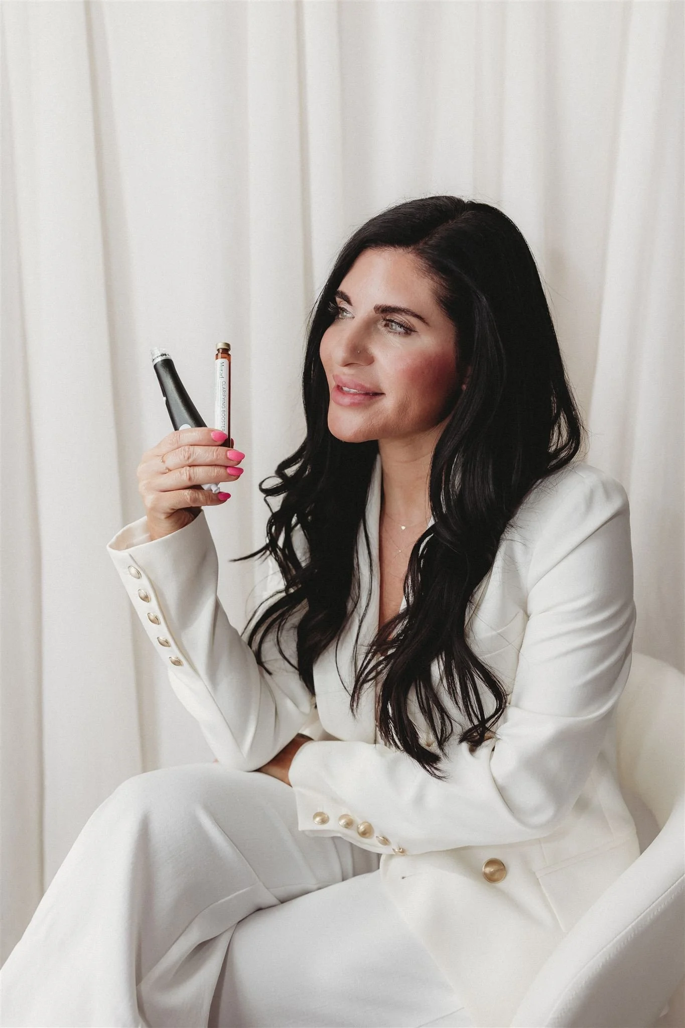 A woman with long dark hair, wearing a white suit with gold buttons, sitting and holding a dental impression device, with a white curtain in the background.