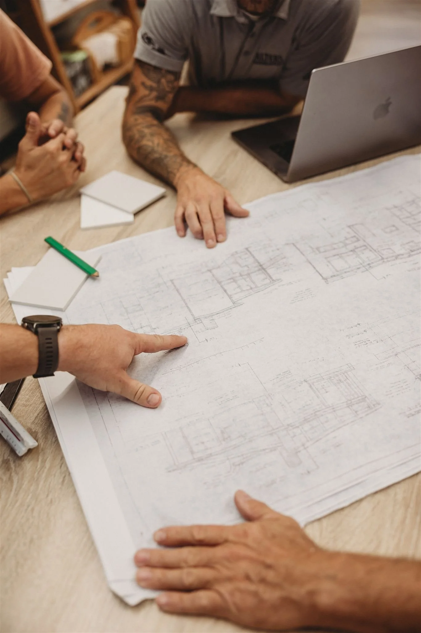 Three people reviewing building blueprints on a table, with a laptop, notepad, and green pen nearby.