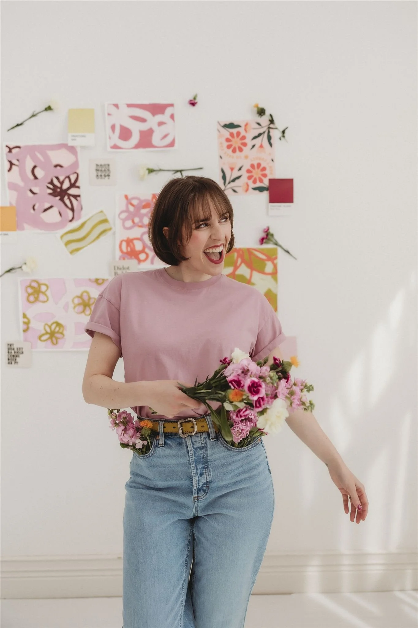 A woman with short brown hair wearing a pink t-shirt and blue jeans, holding a bouquet of pink and white flowers, smiling while standing in front of a white wall decorated with various colorful abstract art pieces and flowers.