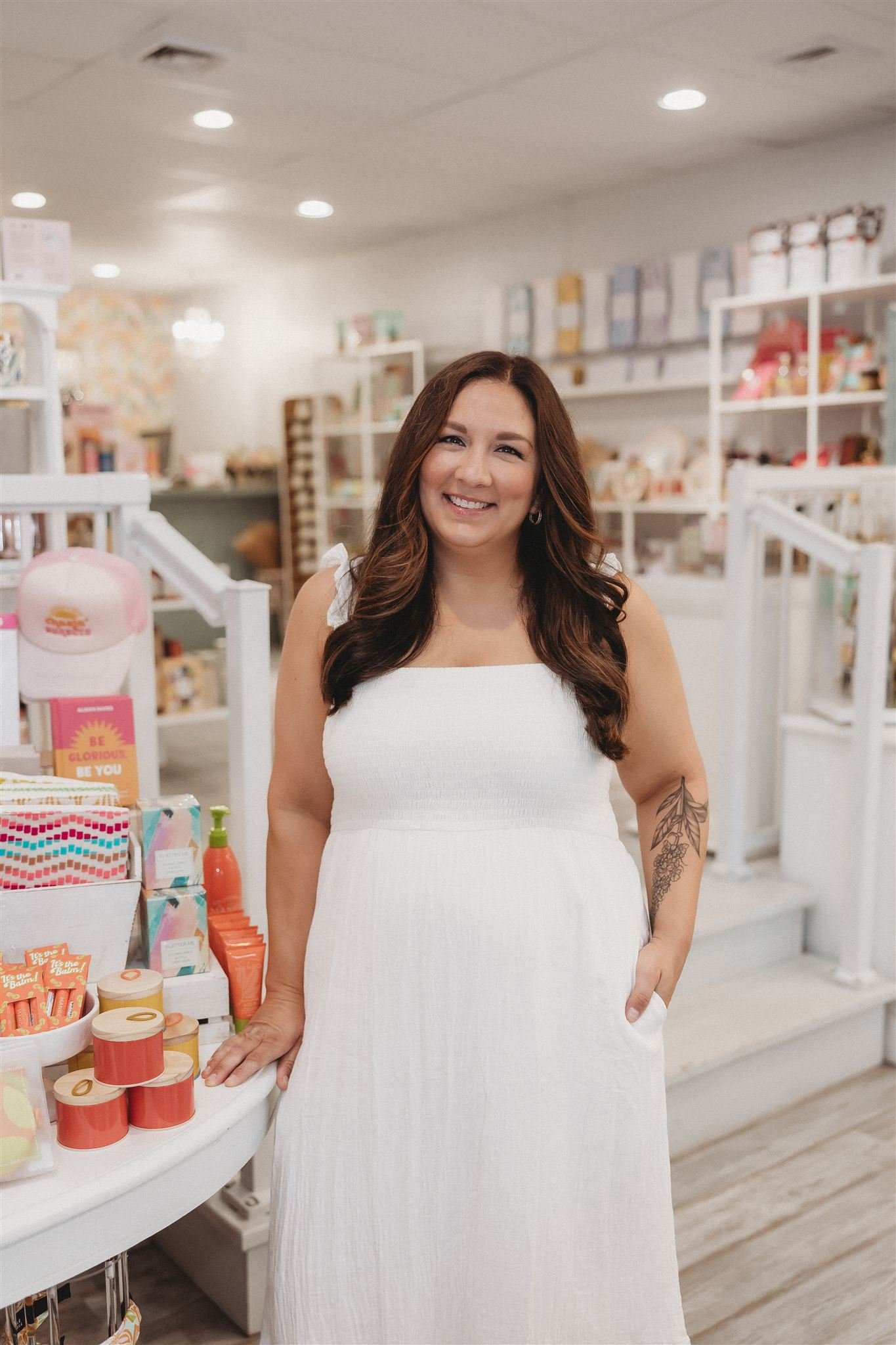 A smiling woman with long brown hair, wearing a white dress, standing inside a store with shelves of products in the background.