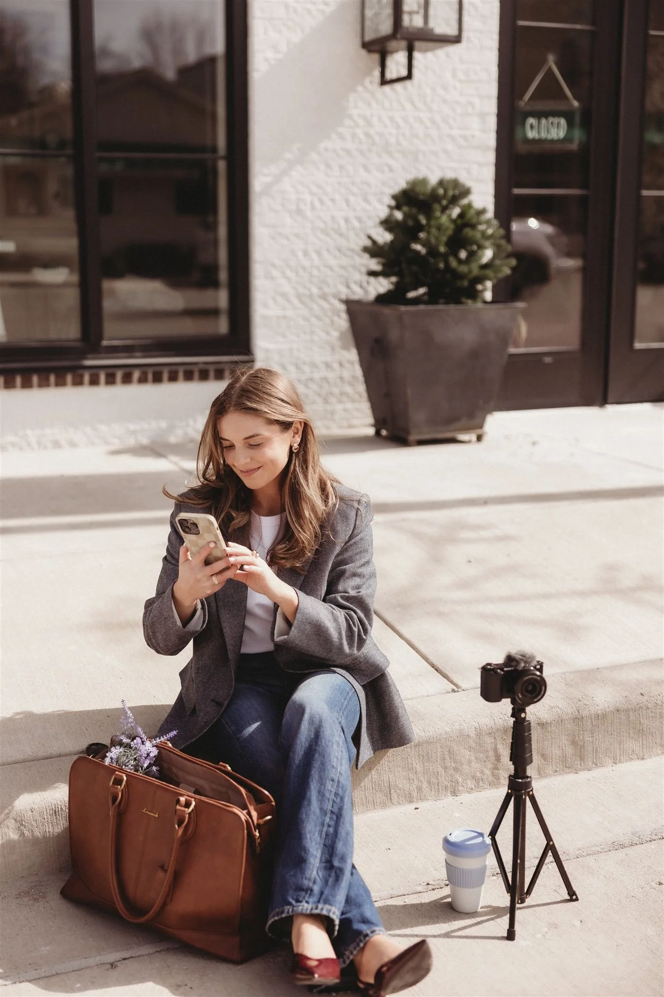 A woman sitting on a sidewalk outside a storefront, looking at her phone, with a camera on a tripod, a coffee cup, a brown bag, and some flowers nearby.