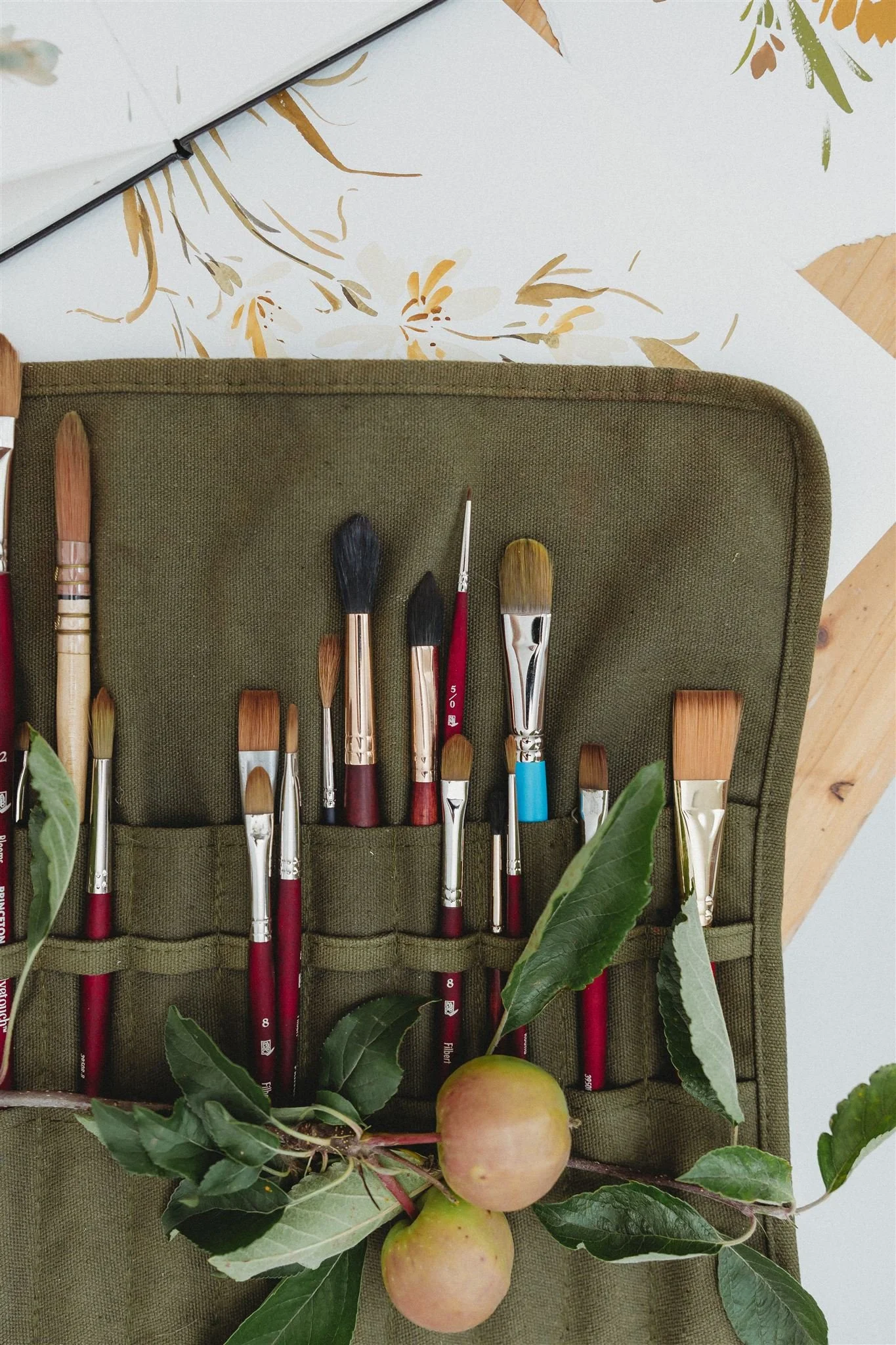 A green pencil case with various paint brushes inside, placed on a table with a branch of apples and leaves on top.