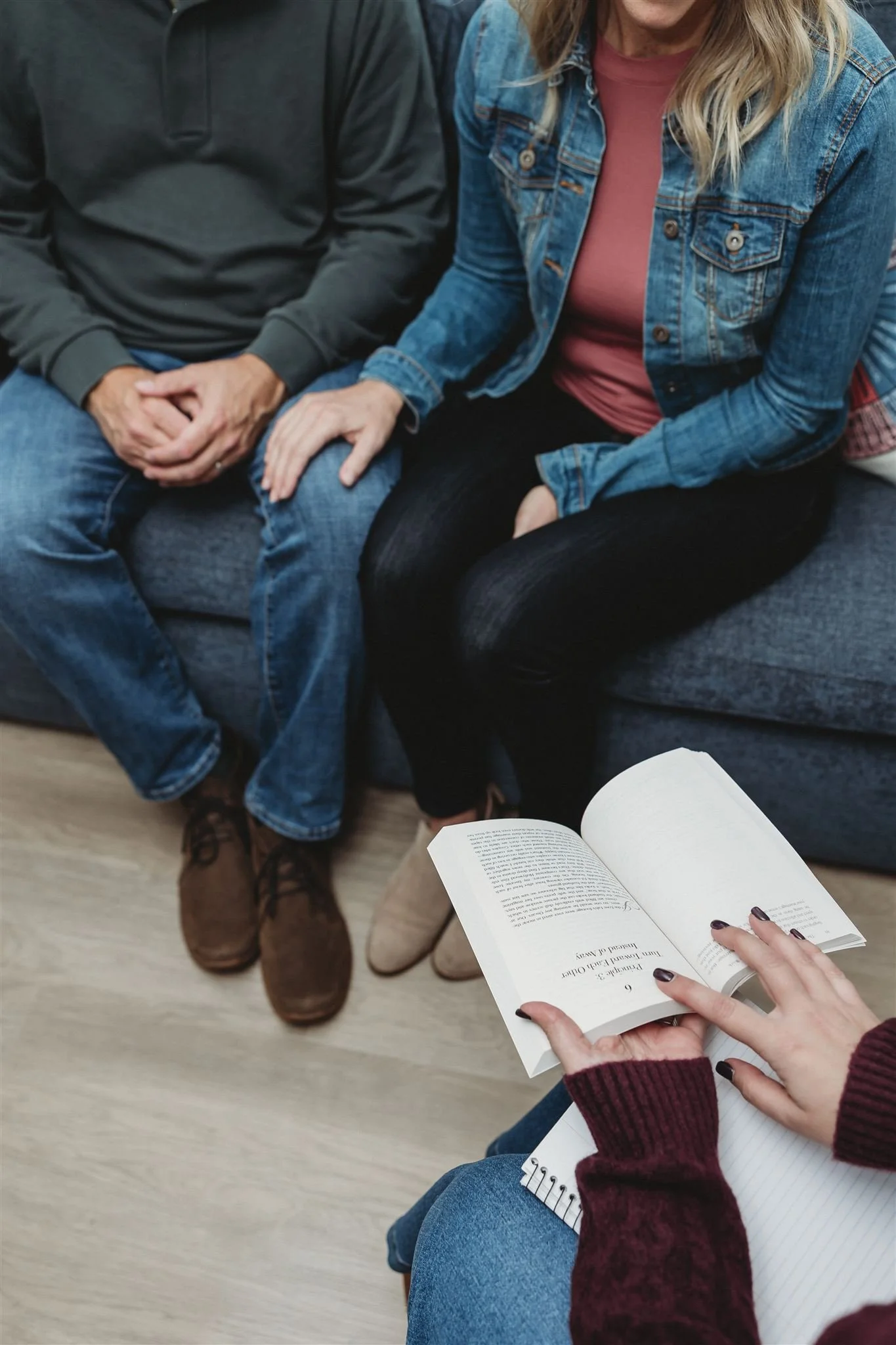 A woman in a red sweater, blue jeans, and dark nail polish pointing to a book held open by another person, with two people sitting nearby on a gray couch, one wearing a black hoodie and jeans, the other in a pink shirt, denim jacket, and black pants, with a wood floor beneath.