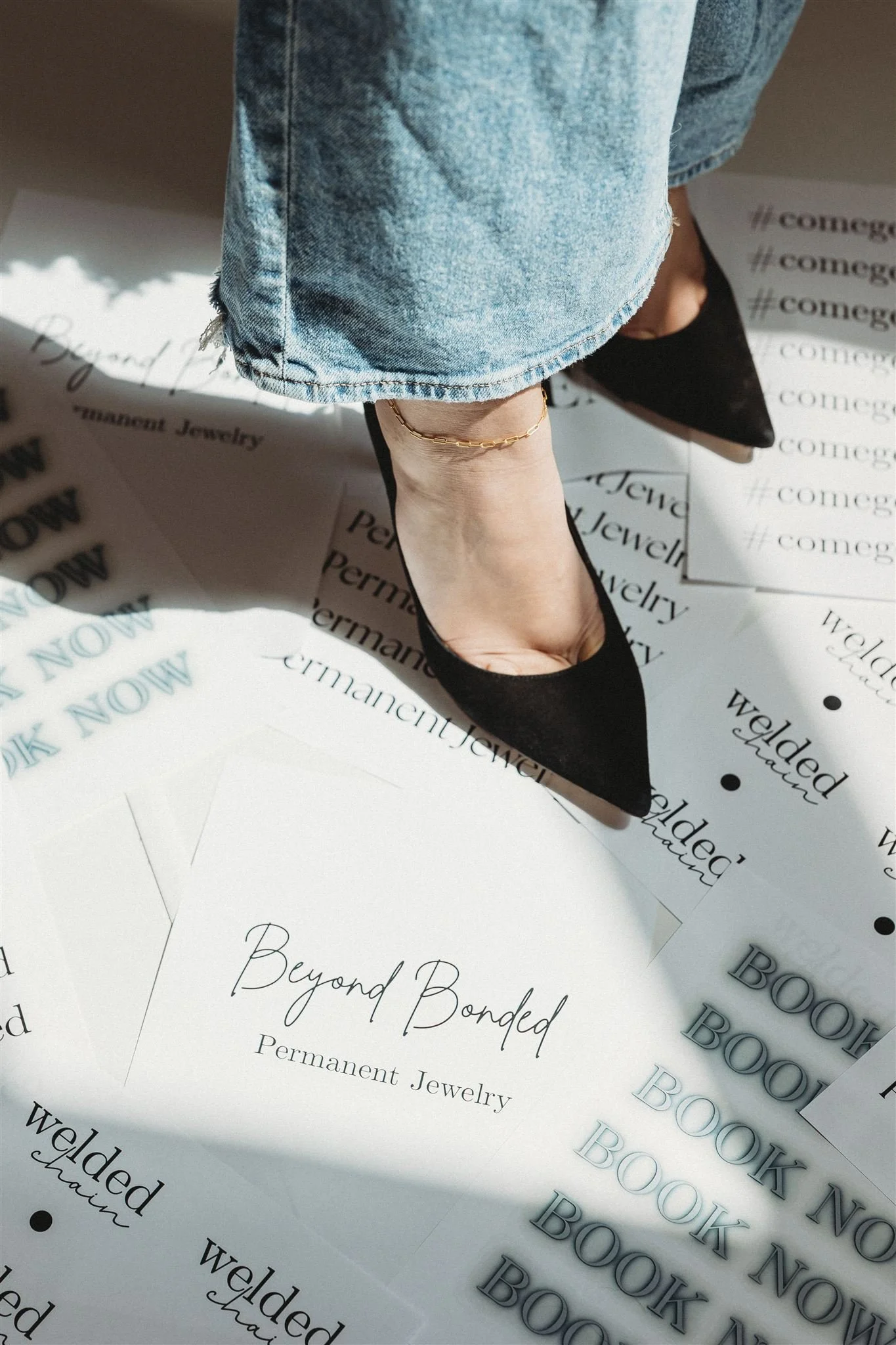A person wearing black pointed-toe heels, a gold ankle bracelet, and ripped blue jeans standing on promotional sheets for Beyond Bonded Permanent Jewelry with the words 'Beyond Bonded' and 'Permanent Jewelry' visible.