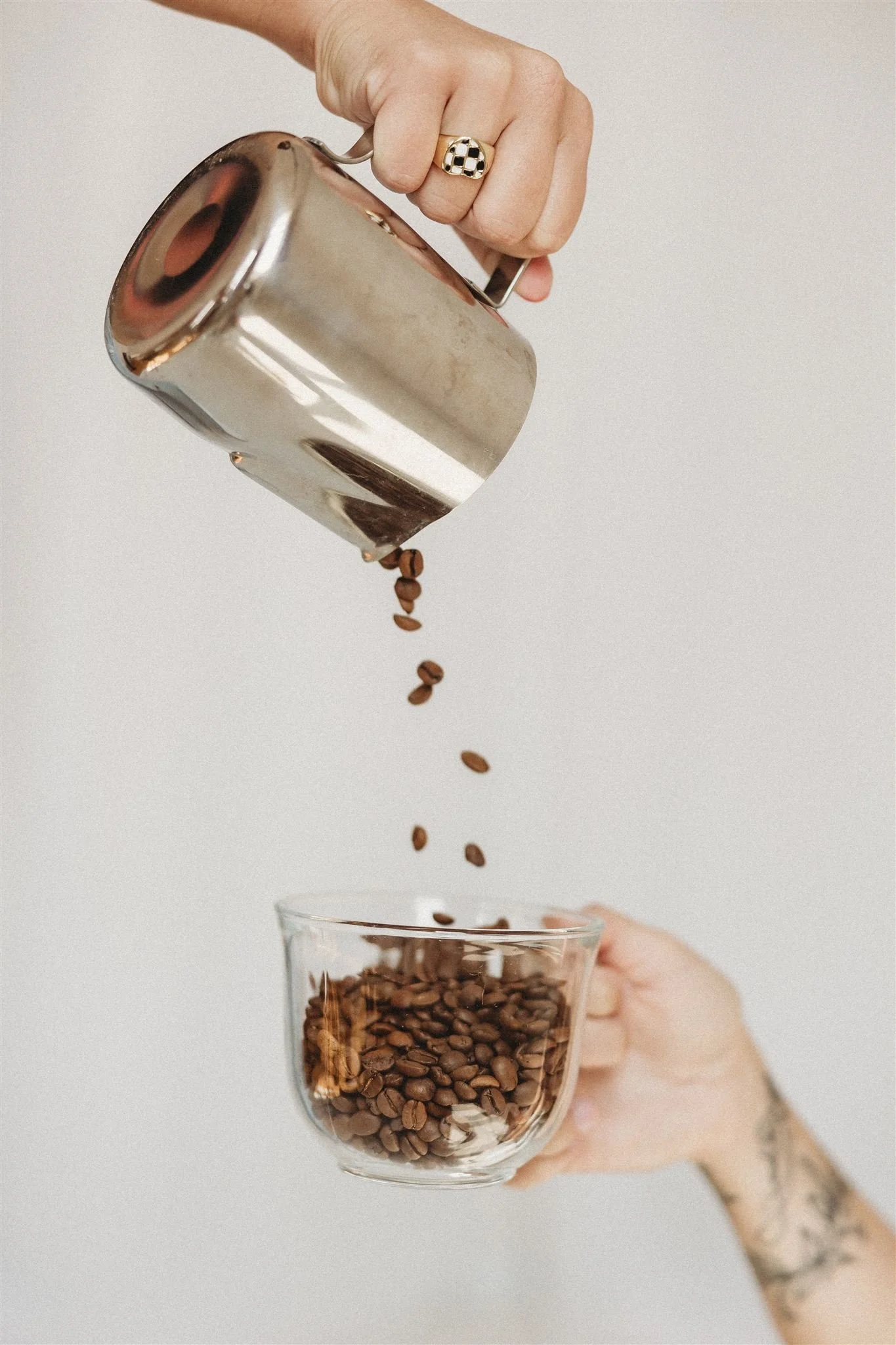 A hand holding a stainless steel coffee pot pouring coffee beans into a clear glass cup, with another hand holding the cup.