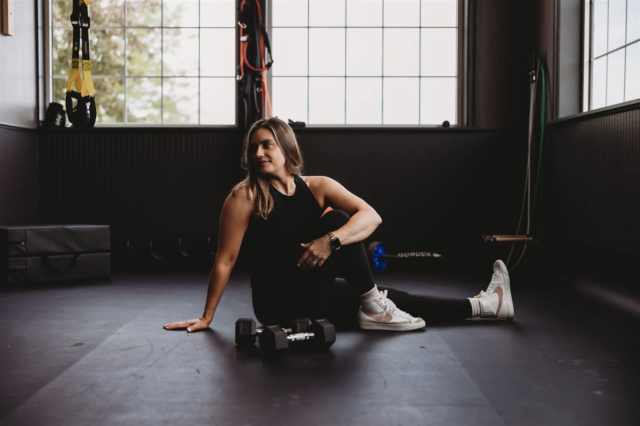 A Vitality Physical Therapy and Wellness physiotherapist sitting confidently on a gym floor beside dumbbells and TRX equipment, embodying their integrated approach to women's health, strength-based rehabilitation, and functional fitness recovery.