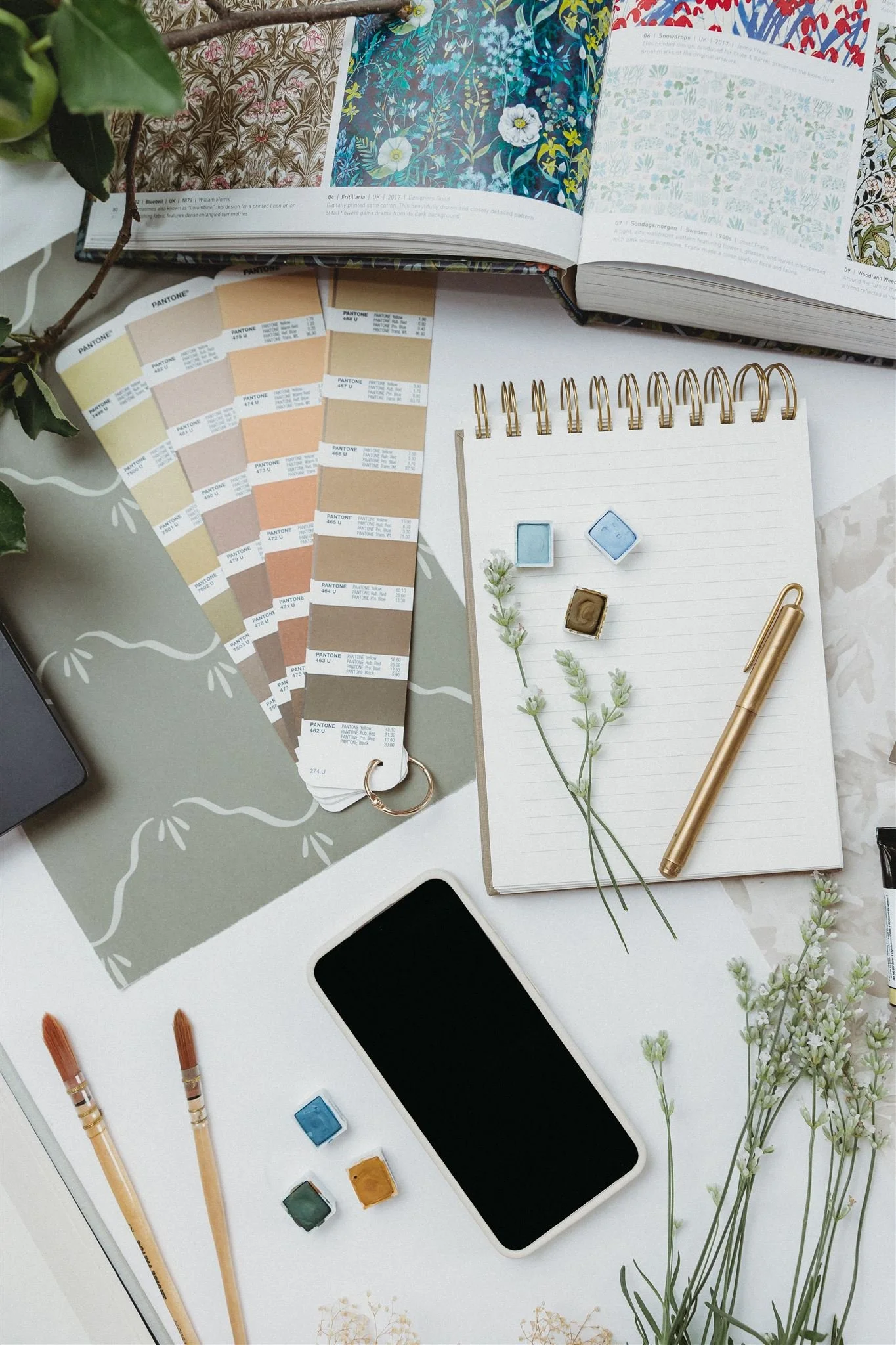 A desk with open design books, paint swatches, watercolor paints, a notebook with sprigs of greenery, a gold pen, paintbrushes, a smartphone, and small potted flowers.