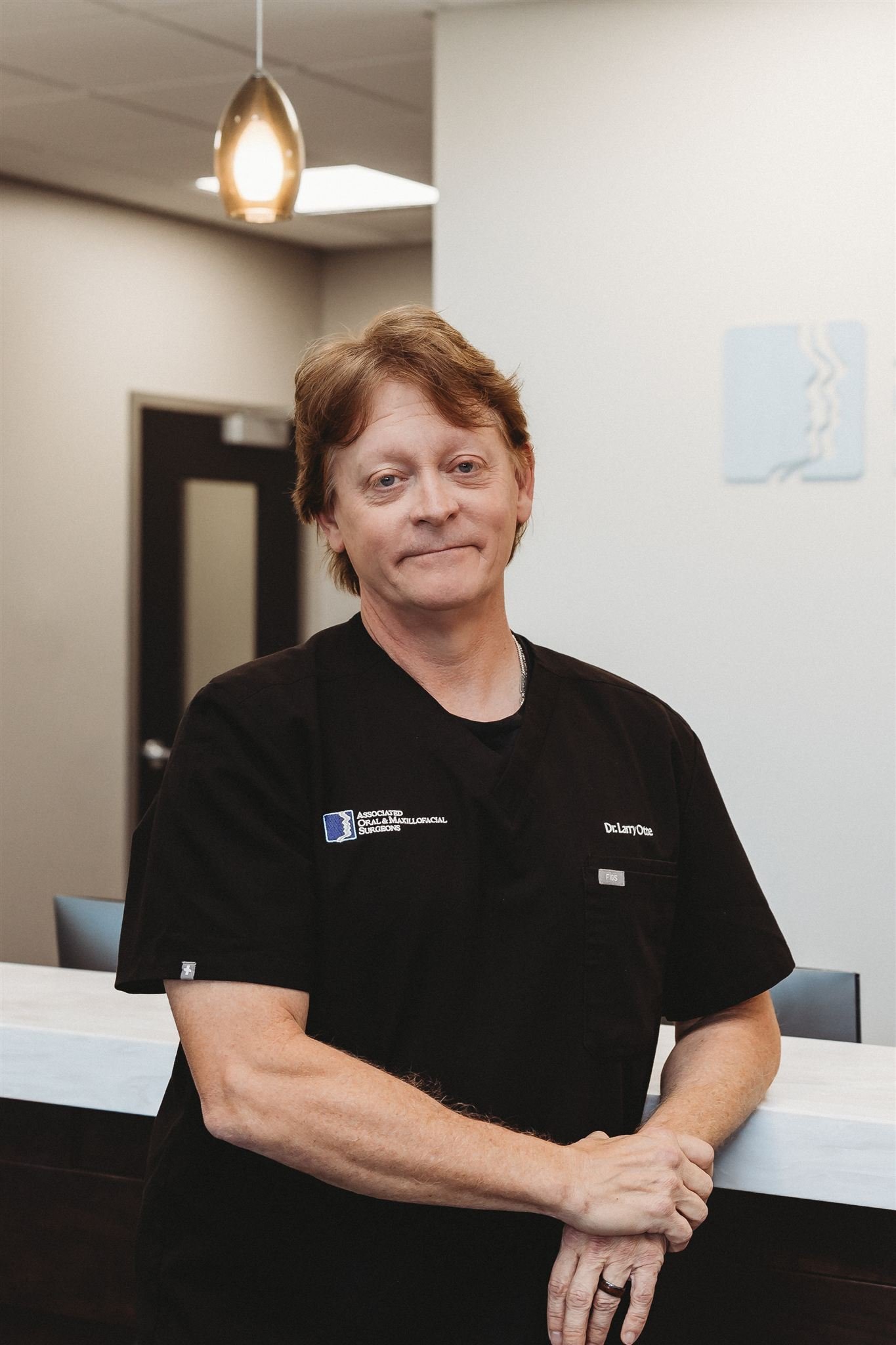 A middle-aged woman with short, light brown hair standing in a medical office or reception area, wearing a black medical scrub with a name badge that reads 'Dr. Larry Otter' and an association patch.