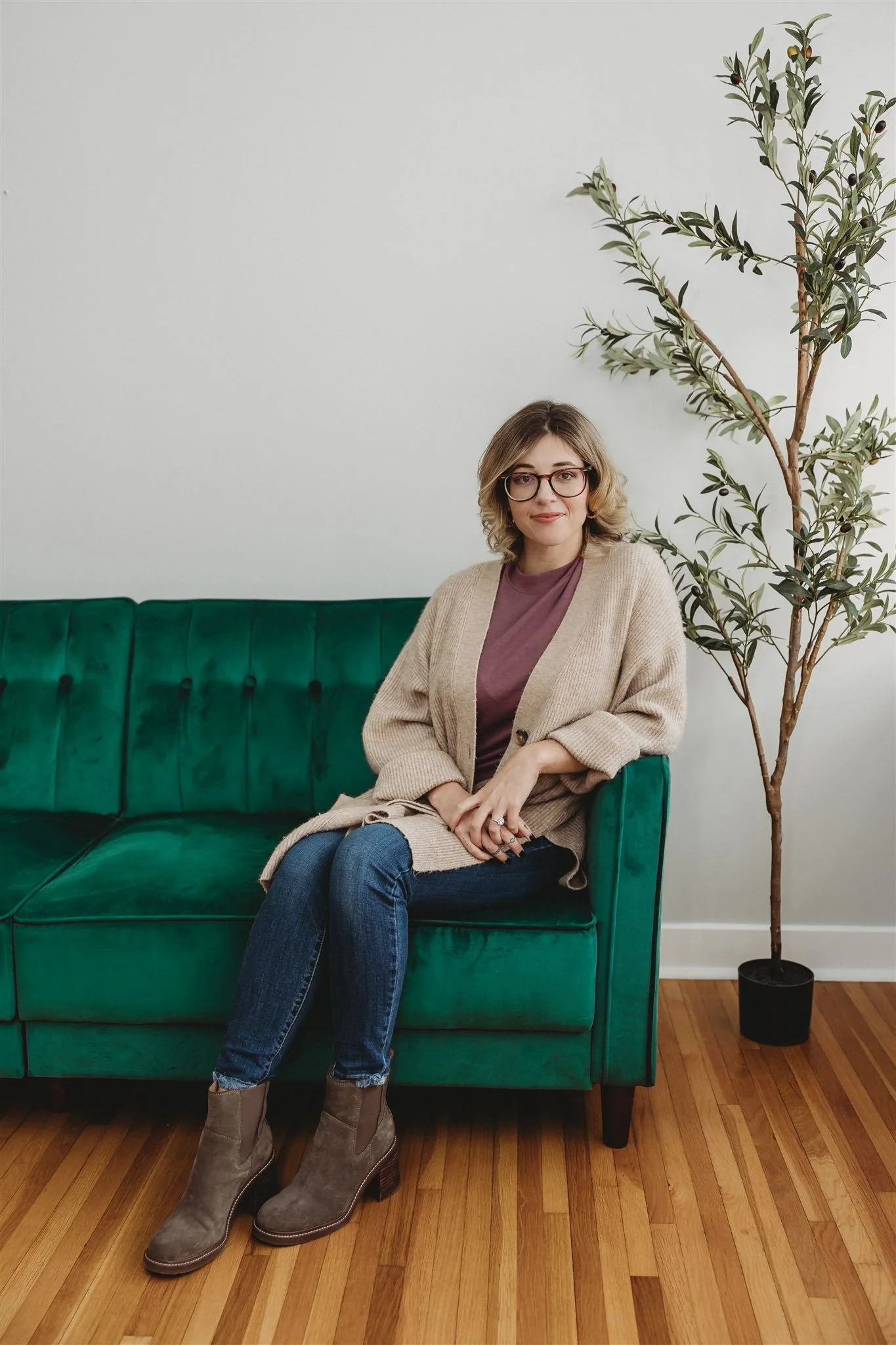 A woman with glasses, short blonde hair, wearing a beige cardigan, purple top, jeans, and brown boots, sitting on a green velvet sofa next to a tall potted plant against a white wall.