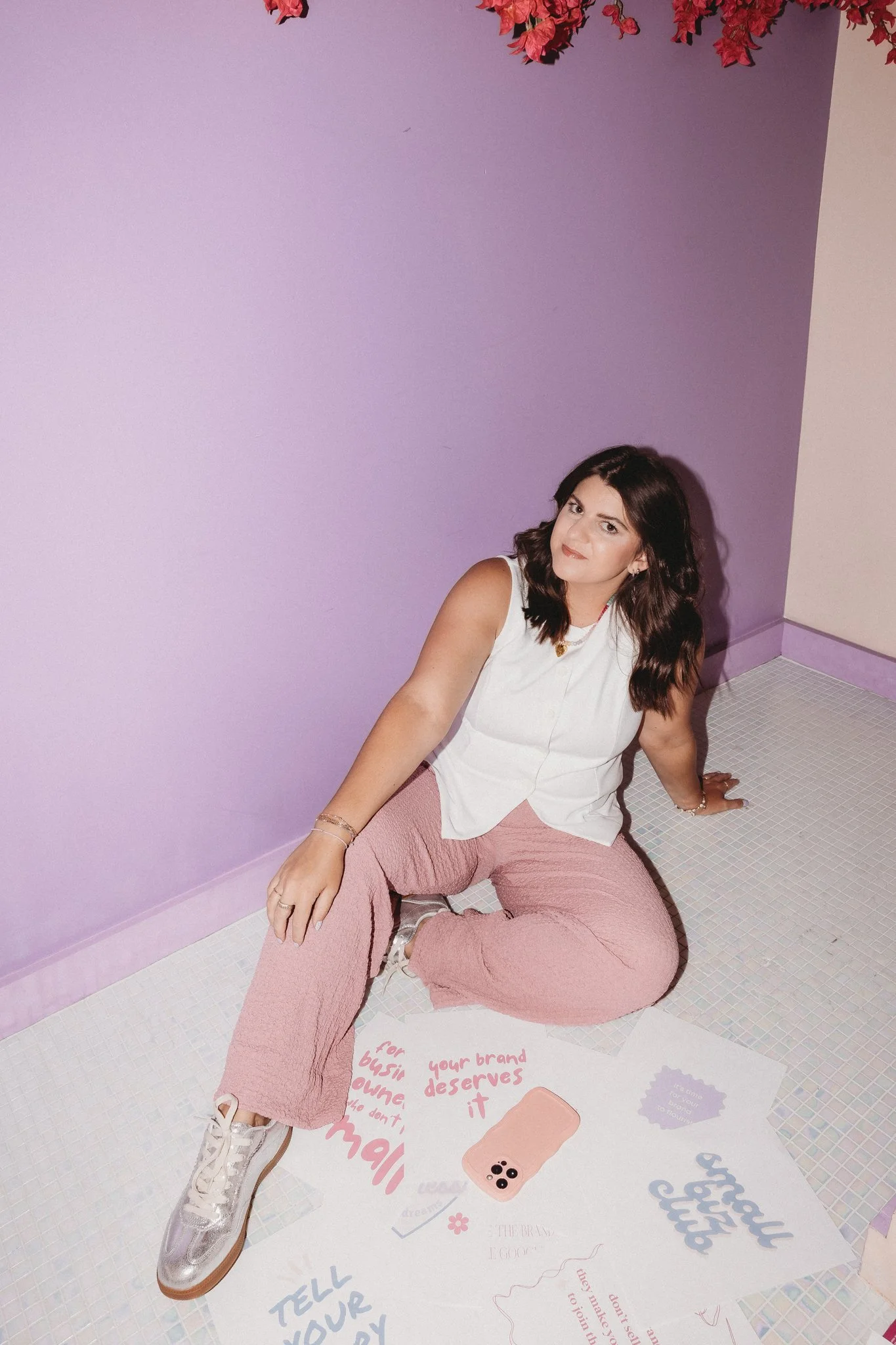 A woman with dark wavy hair and light skin sitting on the floor in a room with purple walls, surrounded by colorful posters and a pink phone case with four camera lenses. She is wearing a white sleeveless top, pink textured pants, and metallic sneakers.