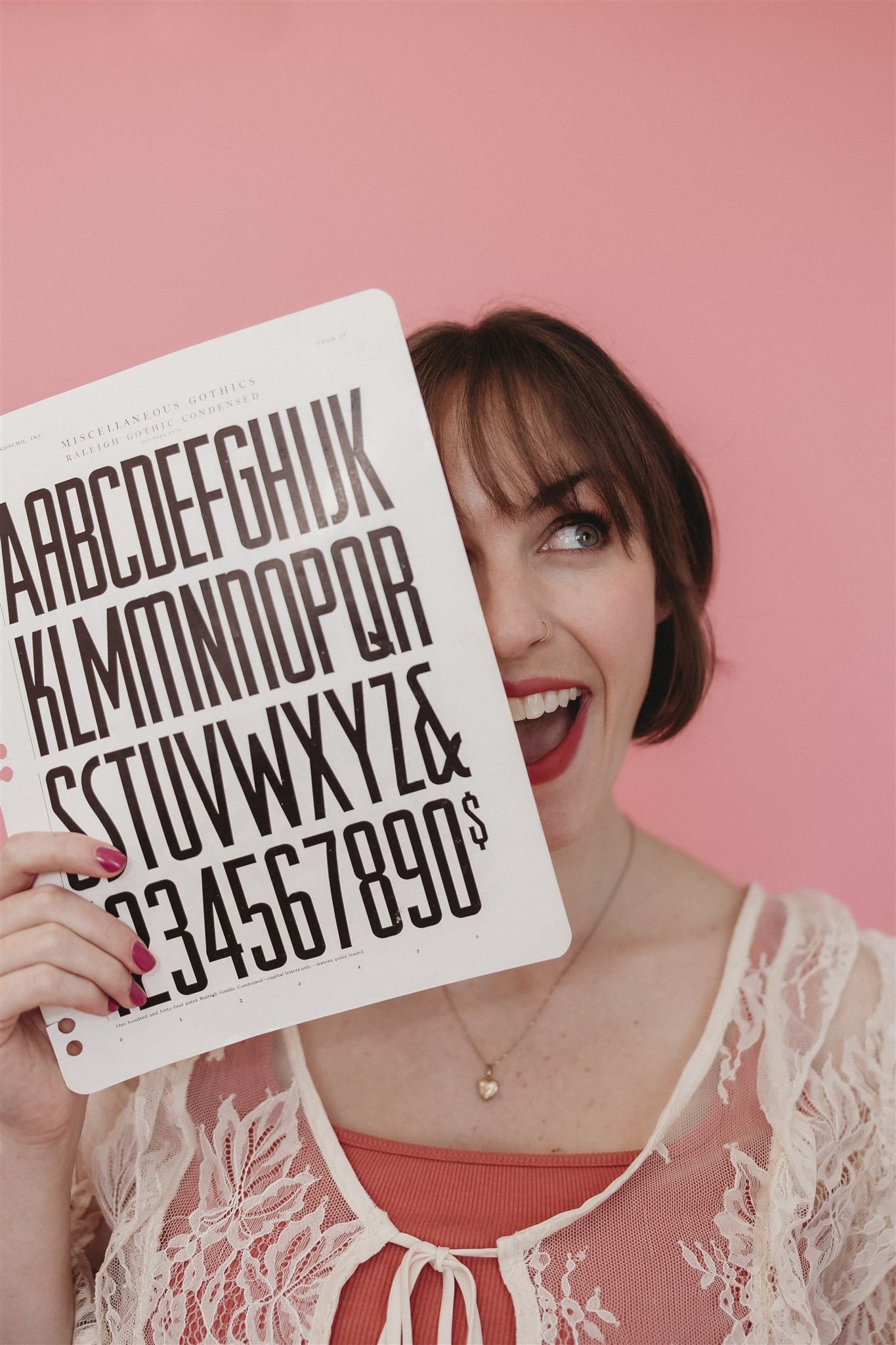 A woman with short brown hair holding a large eye chart in front of her face. She is smiling with a pink background, wearing a lace top and a heart-shaped necklace.