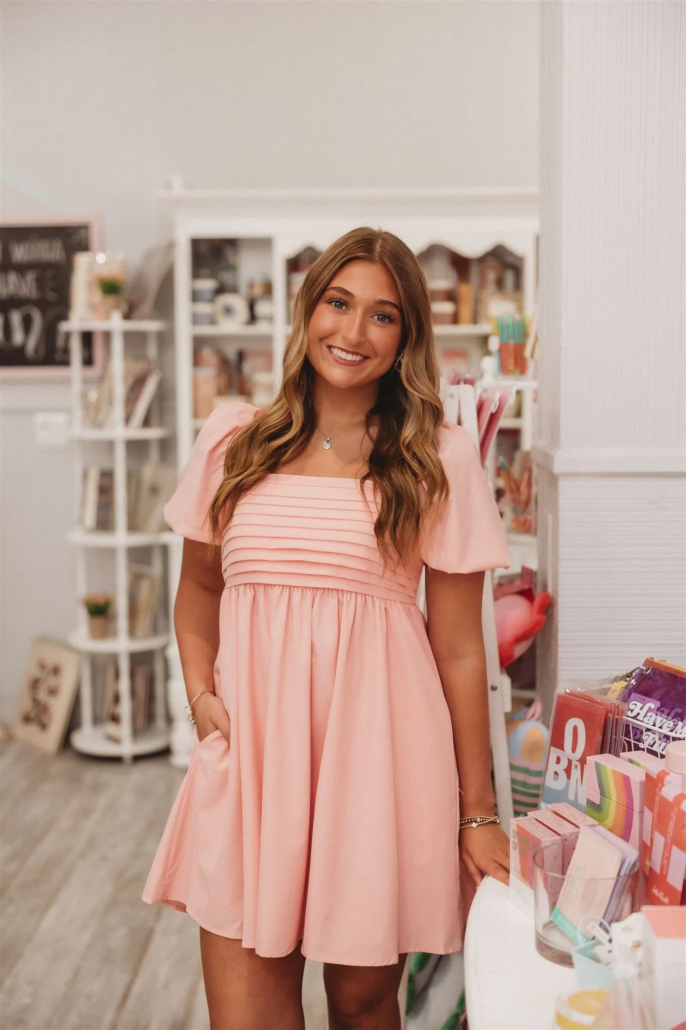 Smiling young woman in pink dress standing inside a store with shelves of books and gifts.