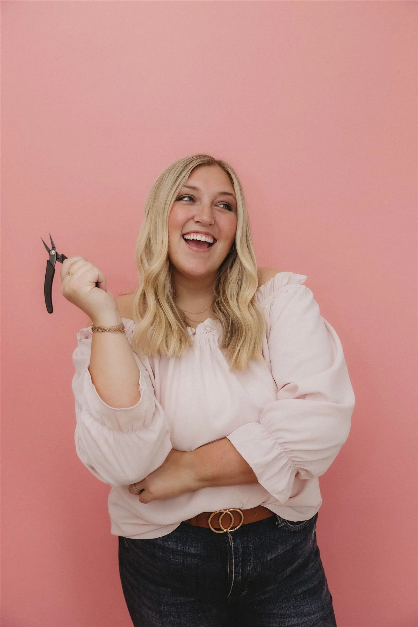 A woman with long blonde hair, wearing a light pink blouse with puffed sleeves, holding a small pair of pliers, smiling and looking to the side, standing against a pink background.