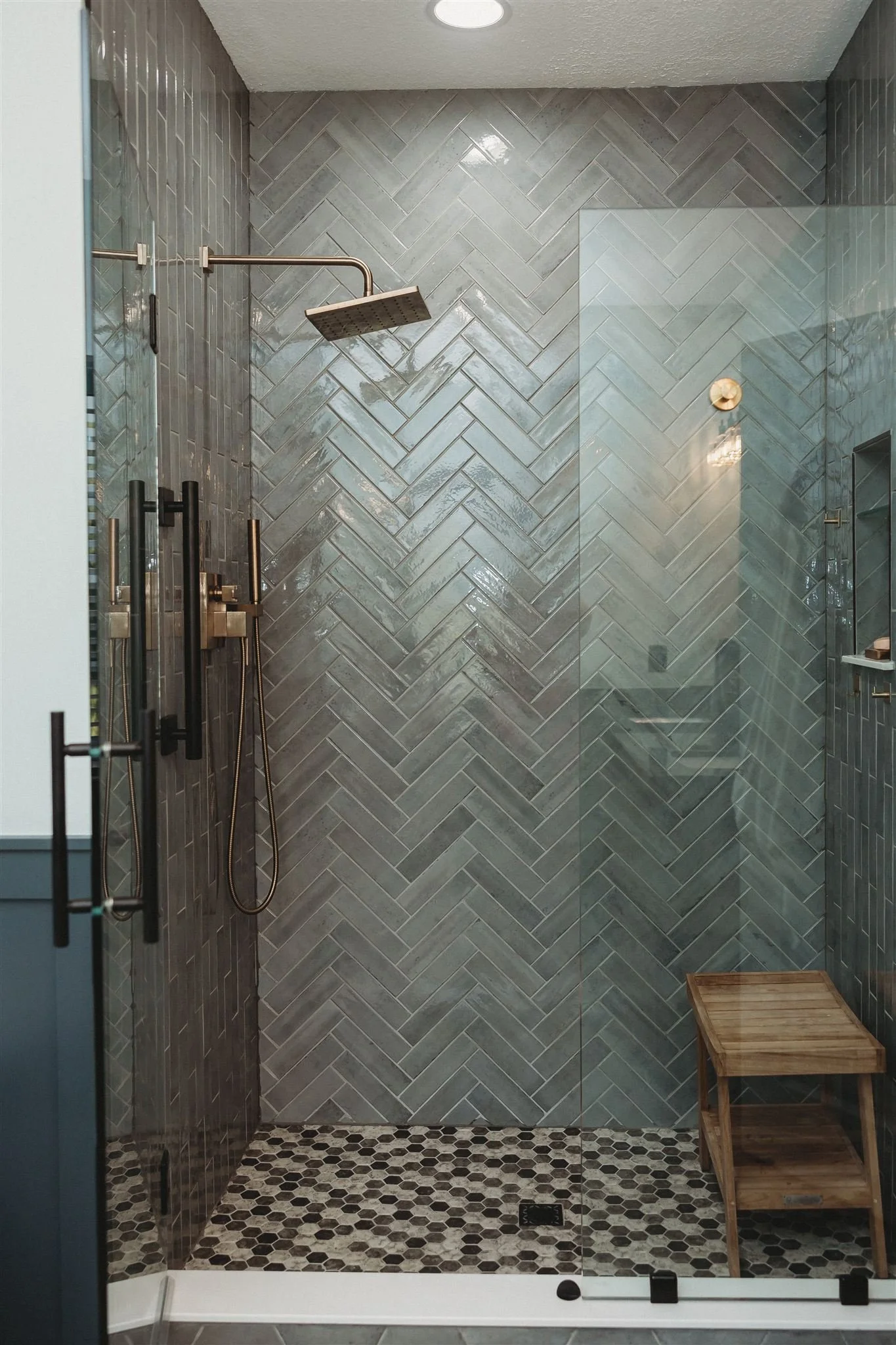Shower area with gray herringbone tile wall, black and white pebble floor, glass door, and a wooden bench.