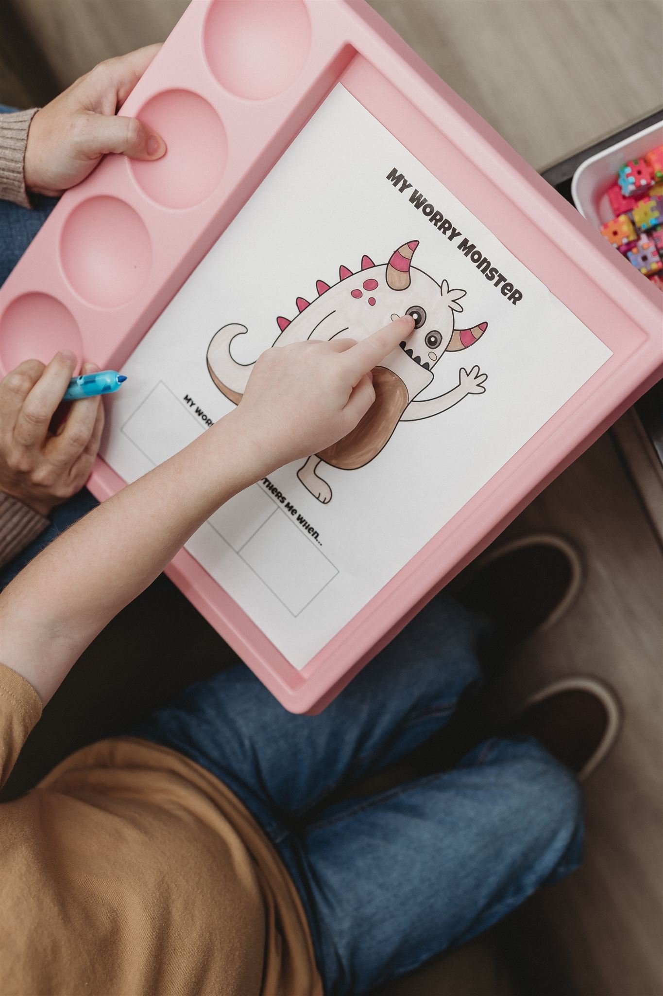 A person coloring a monster drawing on paper titled 'My Worry Monster' with a pink tray containing coloring supplies nearby.