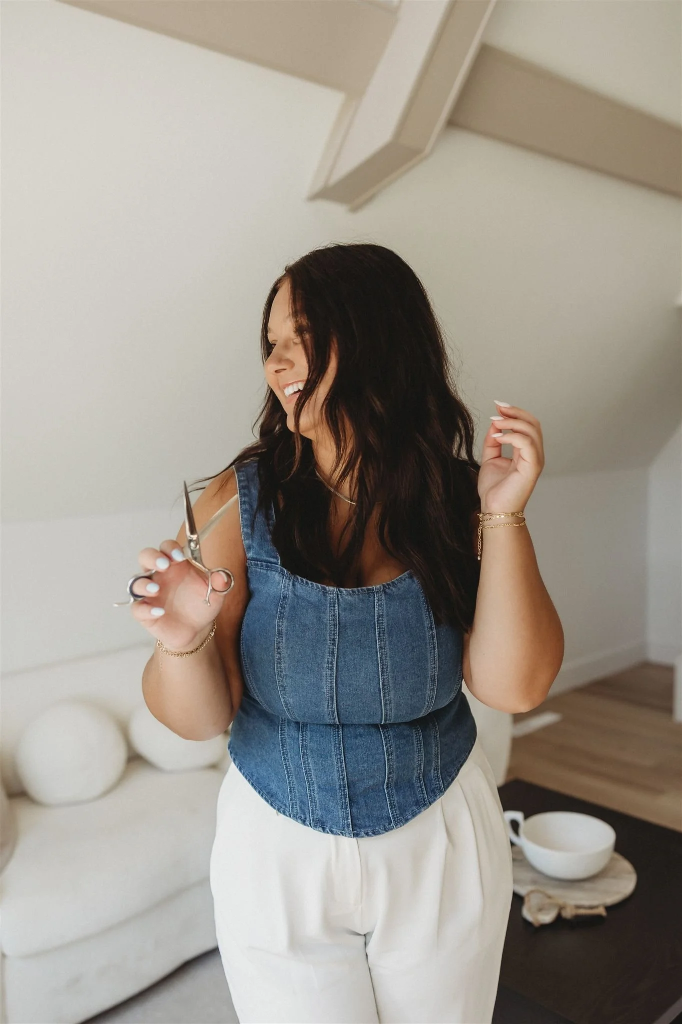 A woman with dark wavy hair, smiling and holding a pair of scissors, standing indoors near a white sofa and a table with a bowl.