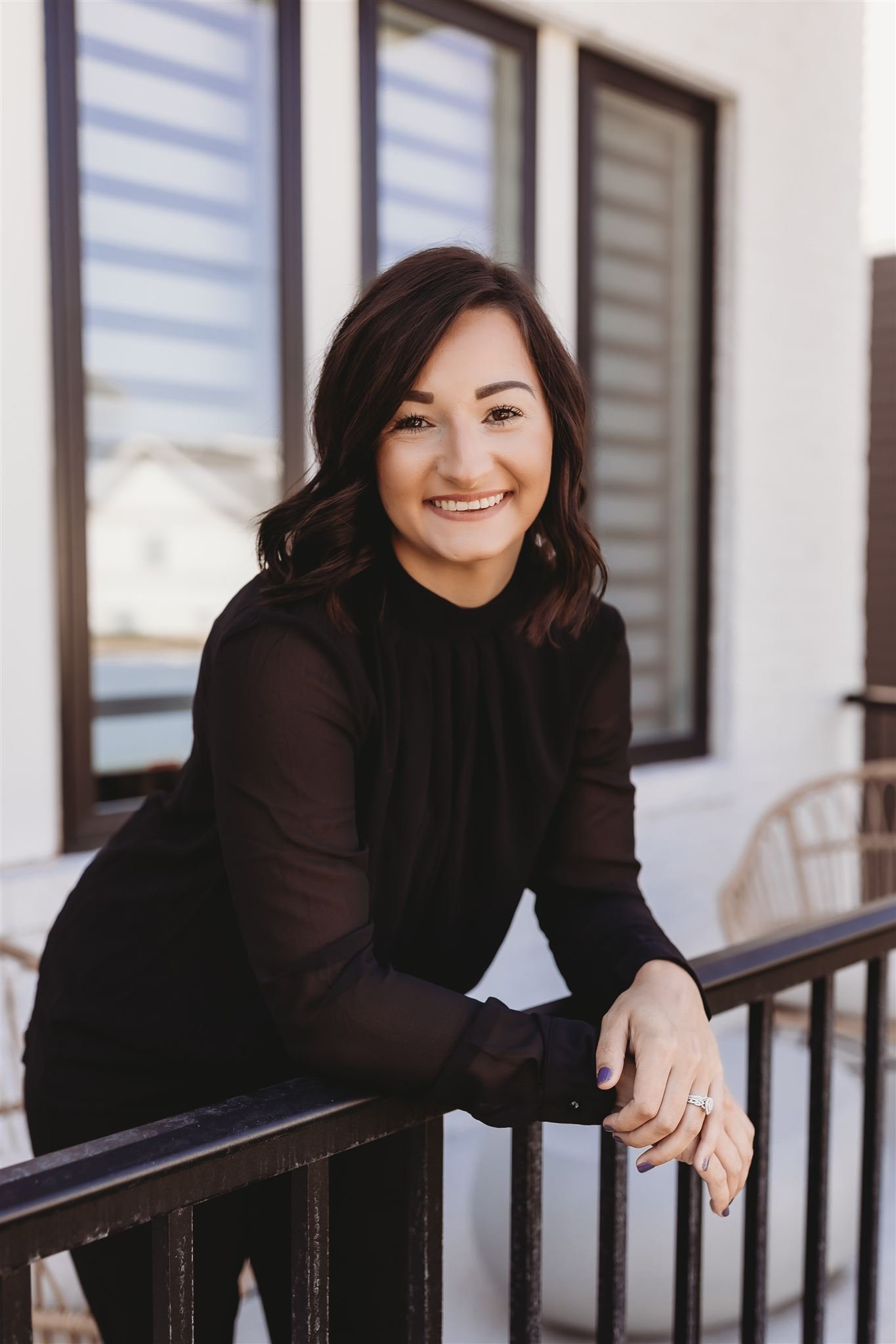 A woman with shoulder-length dark brown hair, wearing a black blouse, smiling while leaning on a black metal railing outside a building with large windows.
