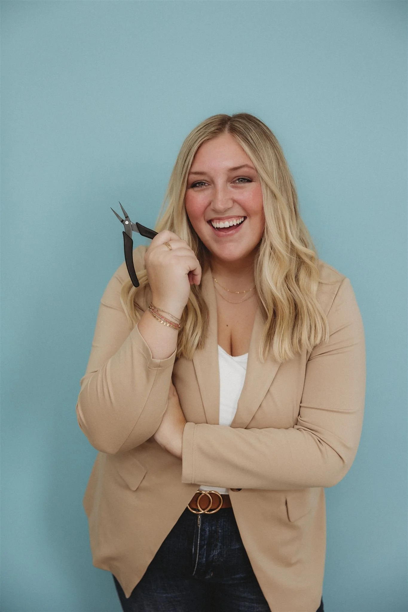 A smiling woman with blonde hair holds a small pair of black manicure scissors near her face against a light blue background. She is wearing a beige blazer over a white top and has layered necklaces and bracelets.